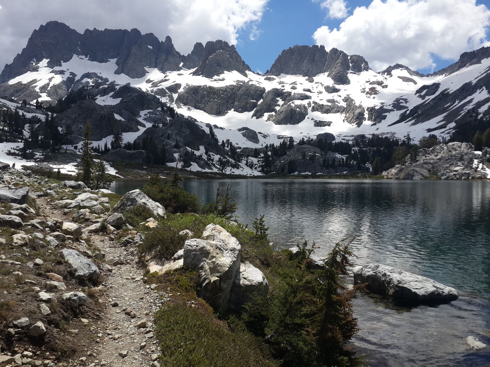 Rocky trail alongside a lake, with snow-capped mountains and cloudy sky in background. - Shadow Creek Trail