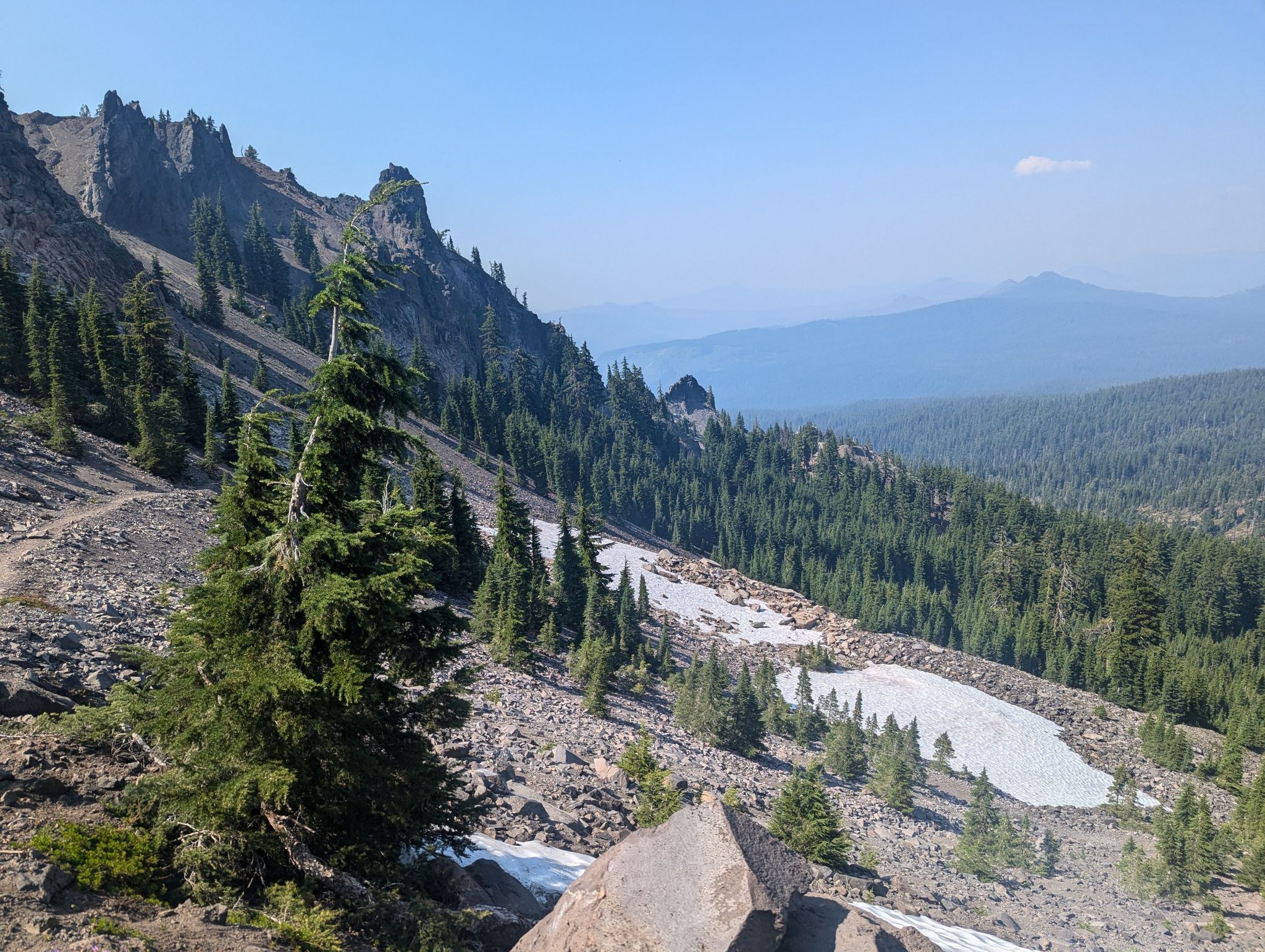 Rocky mountain slope with evergreen trees, snow patches, and distant peaks under clear sky. - Seven Lakes and Devil's Peak Loop