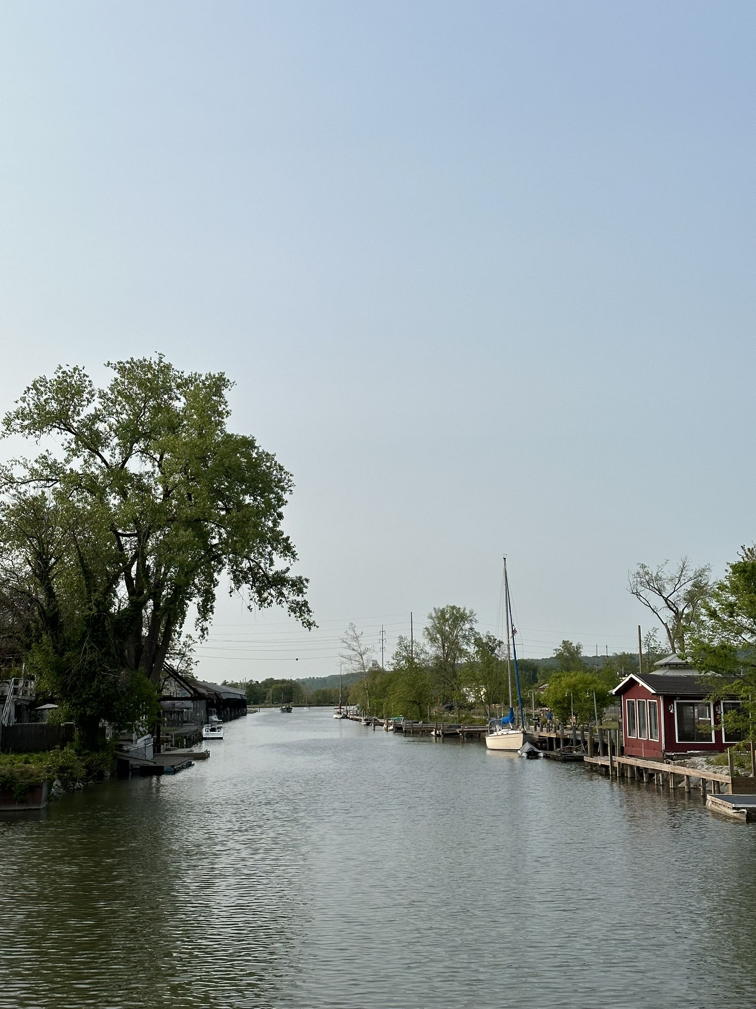 A calm river flows between tree-lined banks with small houses and a sailboat. - Cayuga Waterfront Trail