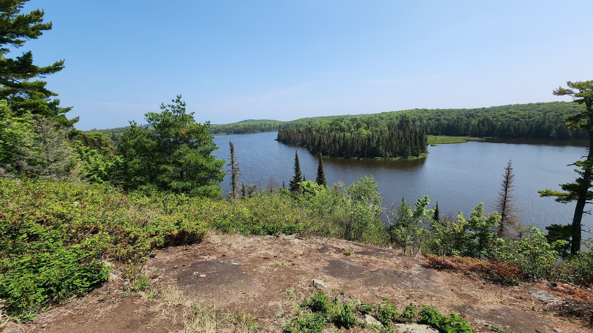 Sunny view of a lake with a forested island from a rocky overlook. - Lake Agnes loop from East Poplar River