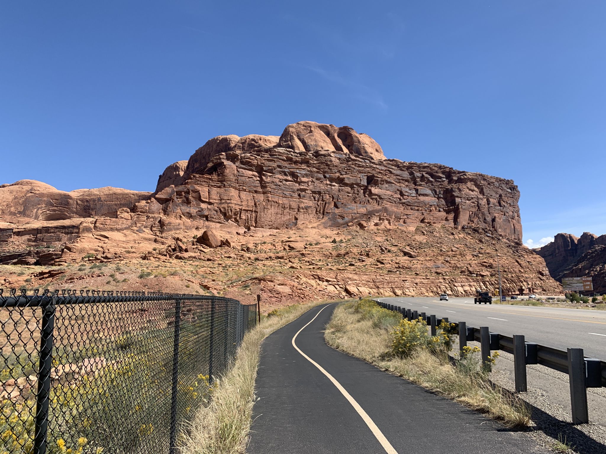 Paved path next to a road, chain link fence, large red rock formation, sunny. - Courthouse Wash Panel Trail
