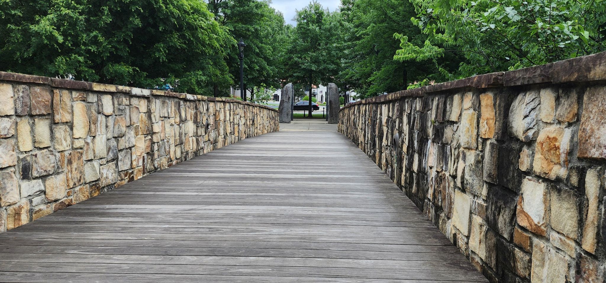 A wooden pedestrian bridge with stone walls leading into a treed area. - Midtown Park – Charlotte Cycles loop from American Legion Memorial Stadium