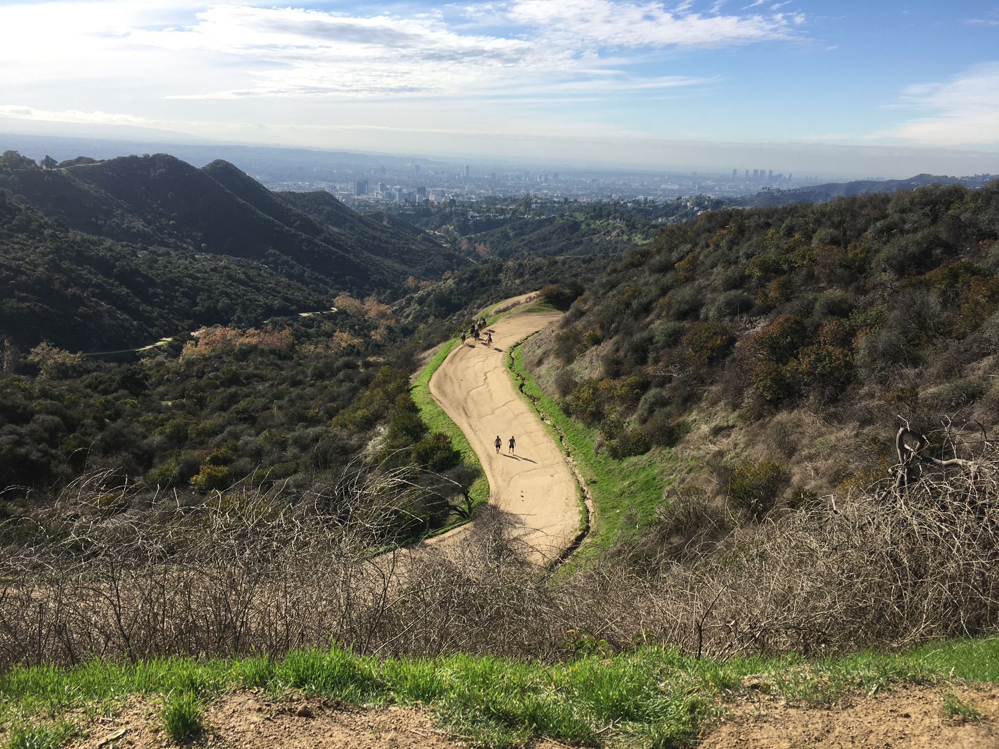 People walk on a winding dirt path through hills with a distant city. - Griffith Observatory – Mount Hollywood Drive loop from Vermont/Beverly