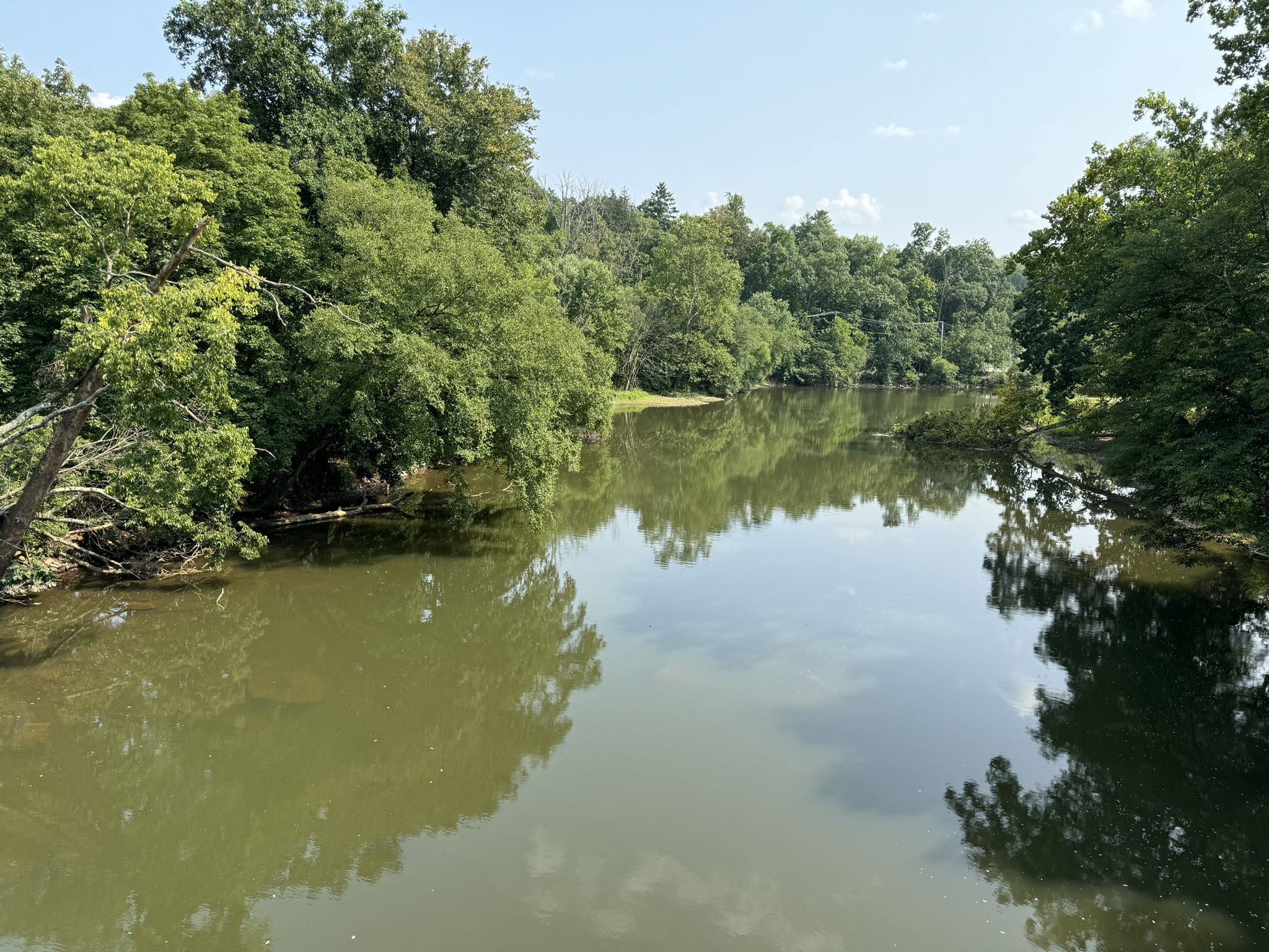 Calm river reflecting green trees and a blue sky with clouds. - Perkiomen Bicycles – Steep Climb on Perkiomen Trail loop from Lower Salford Township