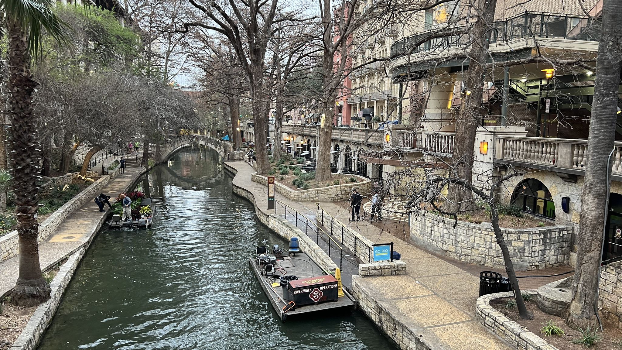 River with boats, stone path, buildings, and bare trees on a cloudy day. - River Walk – Alamo Mills Dam loop from San Antonio
