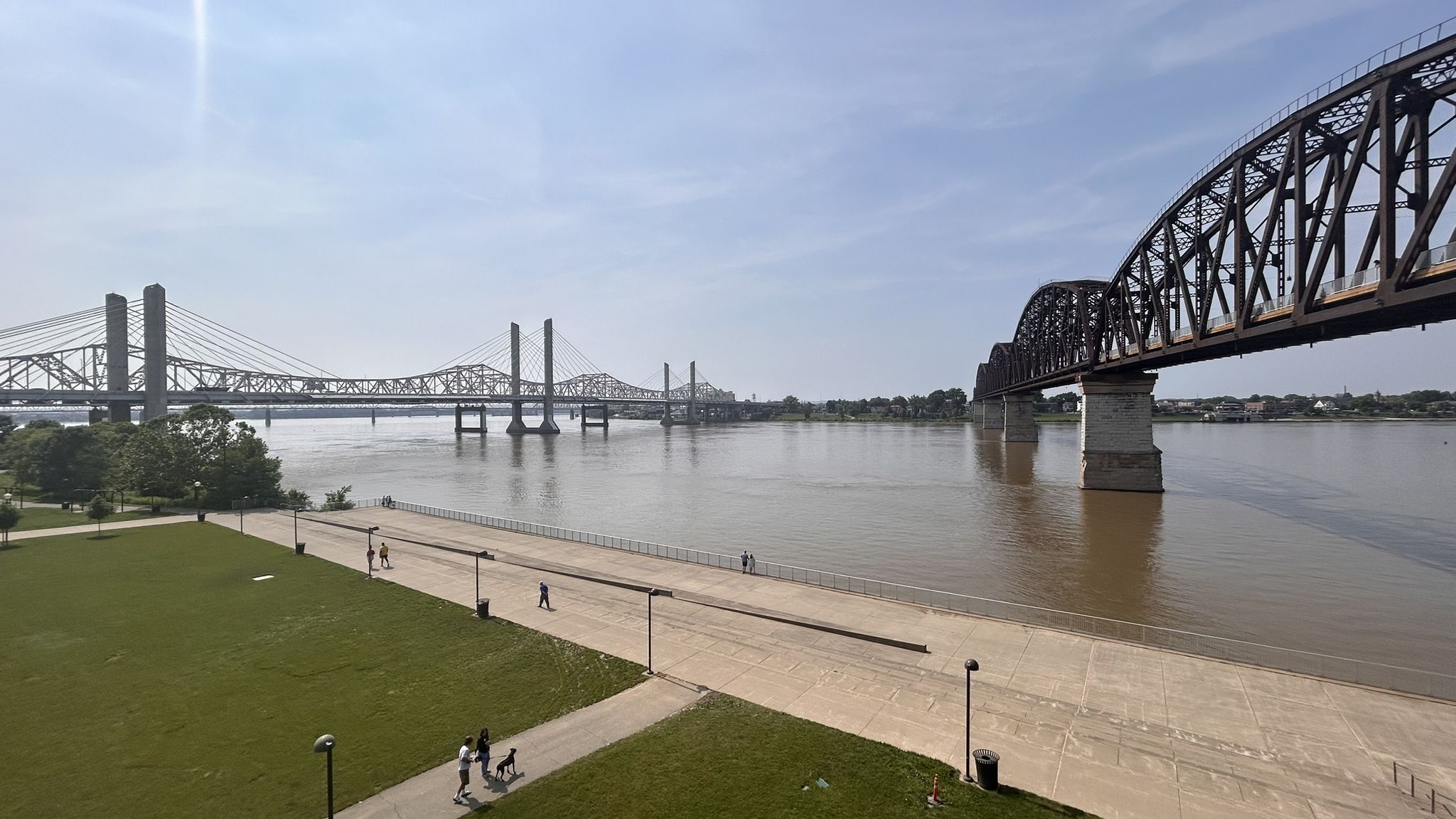 Two bridges span a wide river, with a park and people walking. - Big 4 Walking Bridge