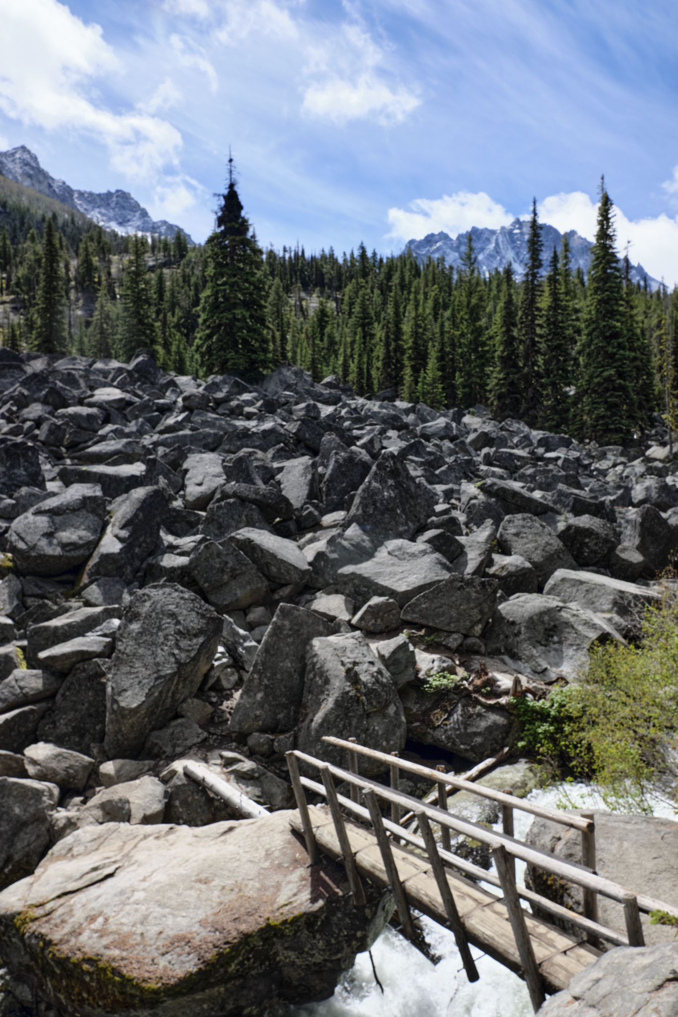 Wooden bridge over rushing water, large rock field, pine forest, and mountains. - Colchuck Lake Trail