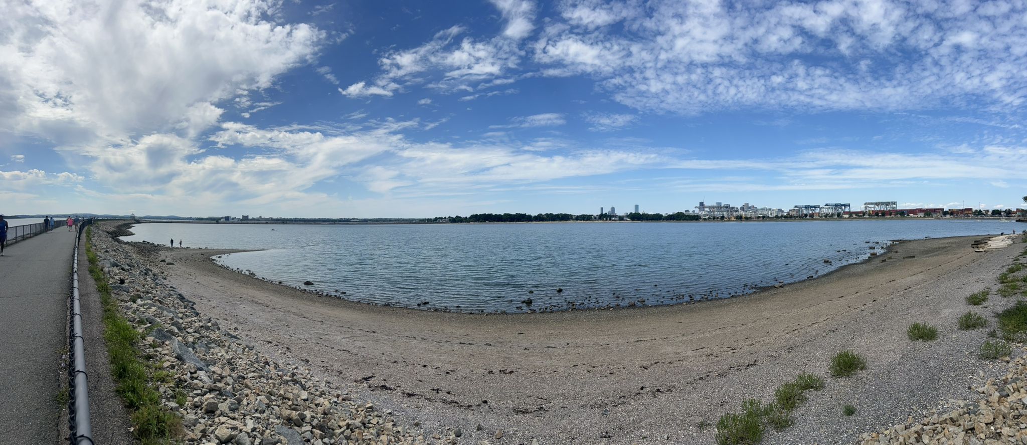 Panoramic view of a wide bay, paved path, rocky shore, and distant city skyline. - Boston Harbor and Castle Island