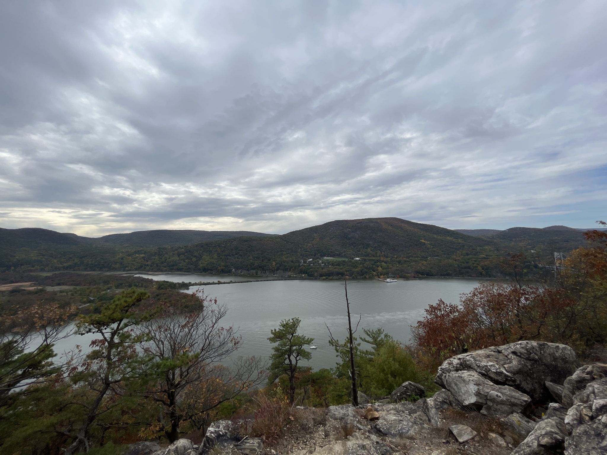 Overcast view of a wide river, forested hills, and rocky foreground. - Anthony's Nose via Camp Smith Trail