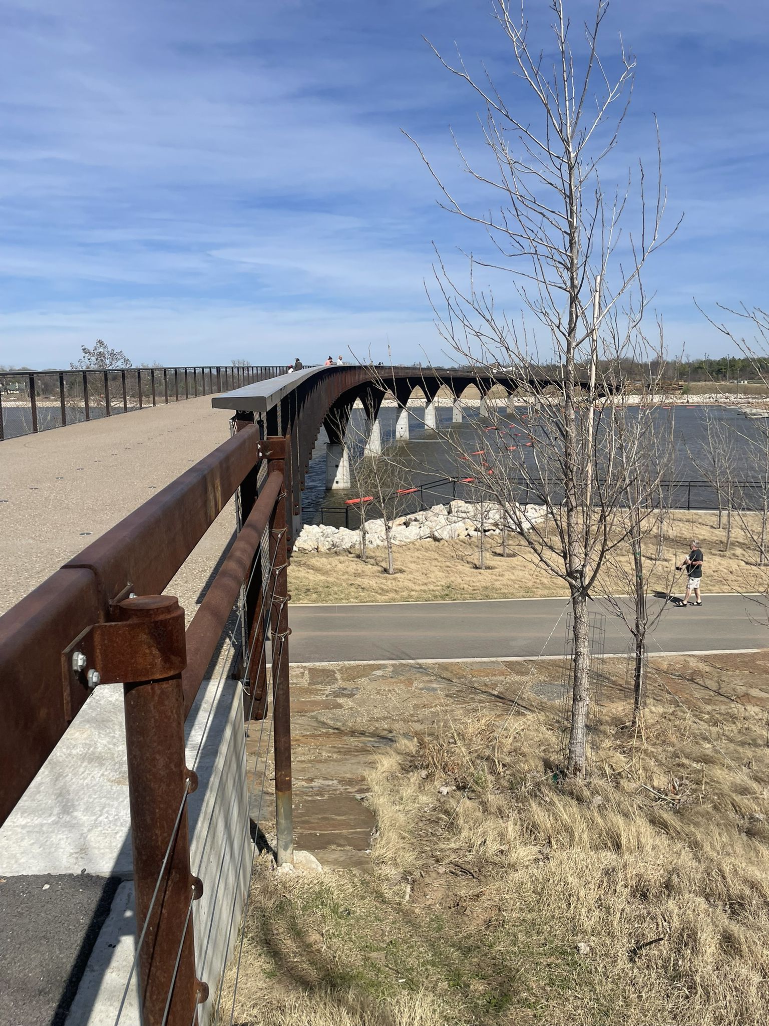 Modern pedestrian bridge with rusty railings extending over water on a clear day. - Gathering Place Park – The Gathering Place loop from All Souls Unitarian Church