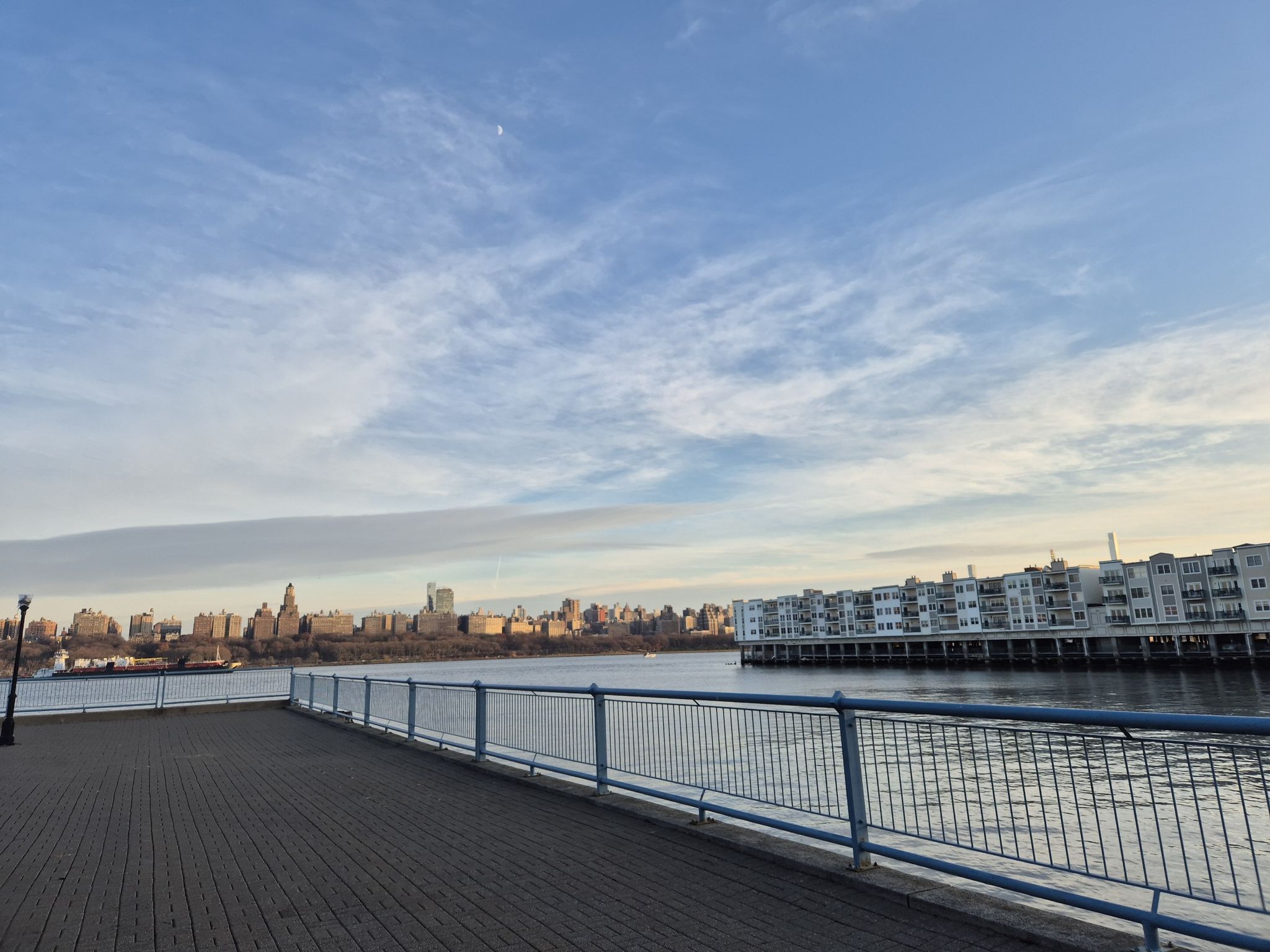Paved promenade next to a river with distant city and waterside buildings. - Port Imperial Waterfront loop from Harborside Financial Center