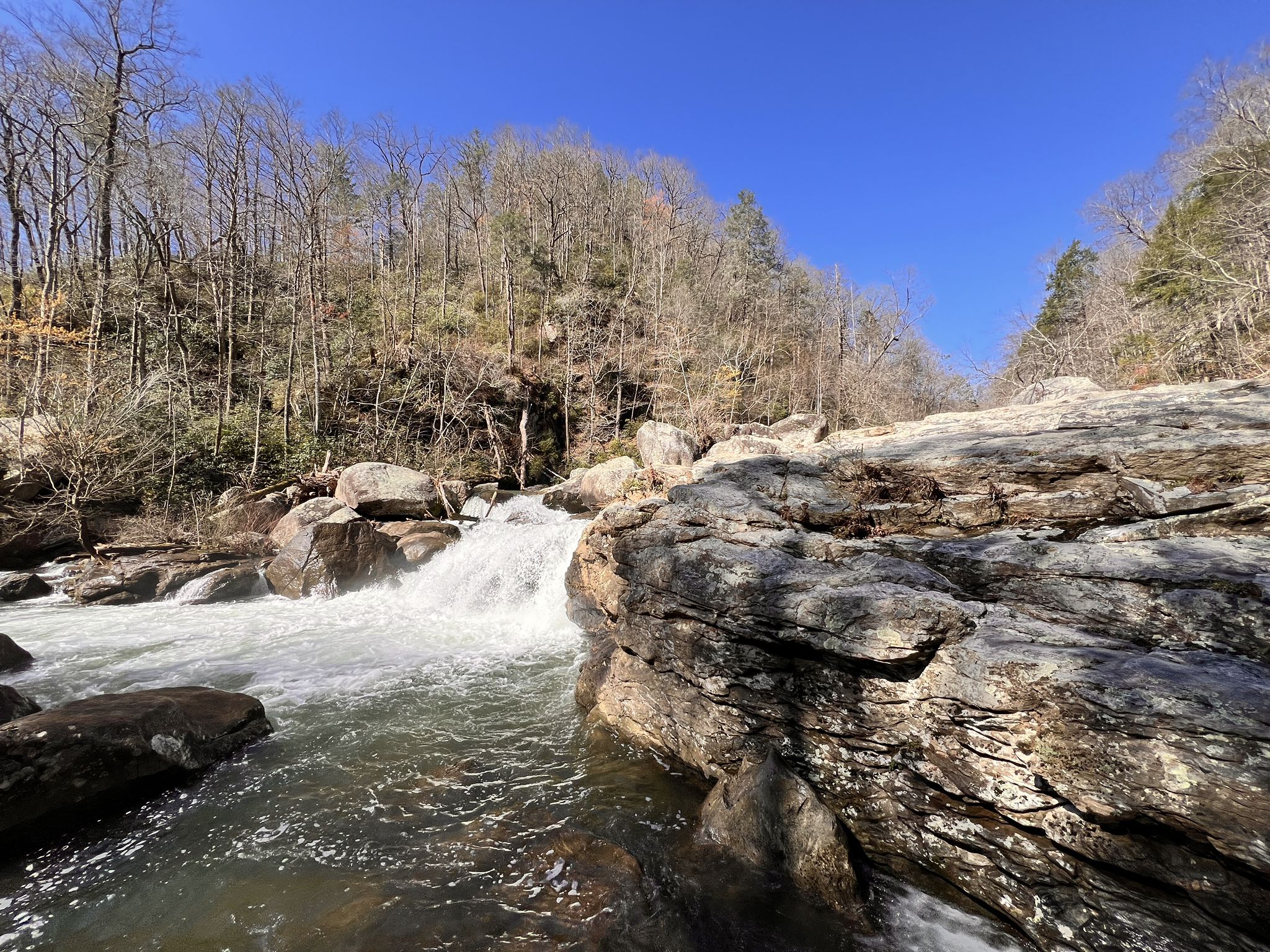A bright waterfall cascades over rocks in a bare winter forest under blue sky. - Pulliam Creek Trail