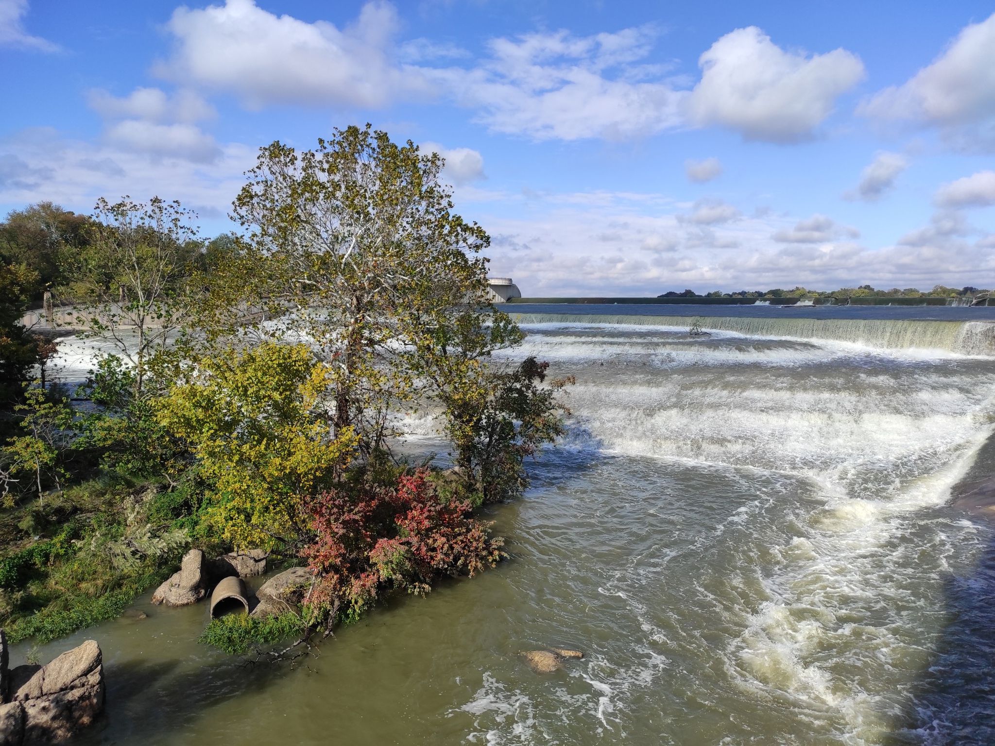 Wide river with a dam and waterfall, framed by vibrant trees. - White Rock Lake Trail