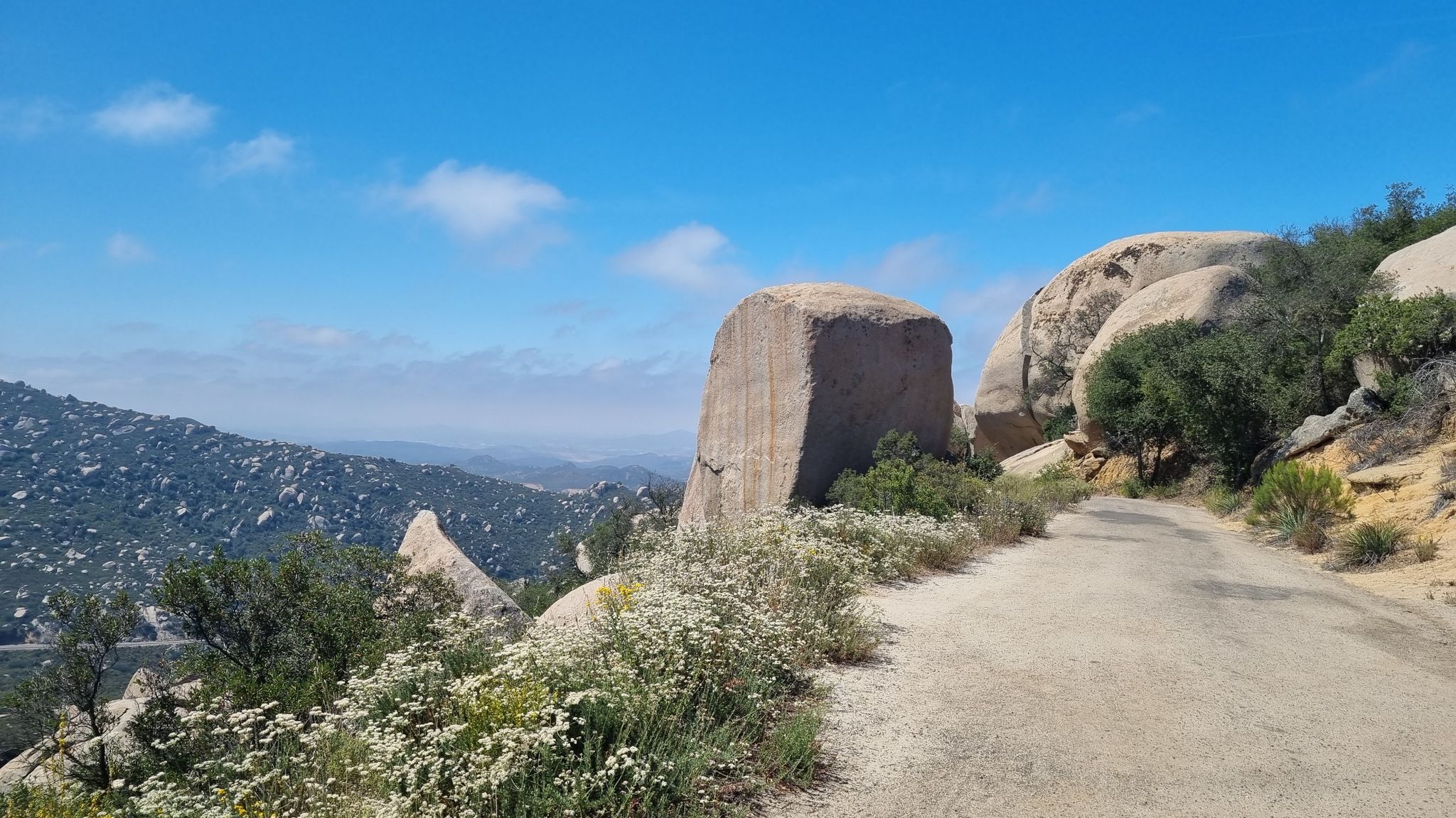 Dirt road winds past large boulders, distant mountains under a blue sky. - Potato Chip Rock via Mount Woodson Road