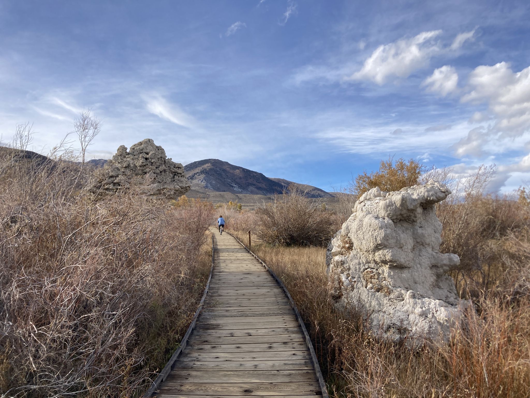 A person walks on a wooden boardwalk through dry brush and rock formations. - Mono Lake Boardwalk Trail