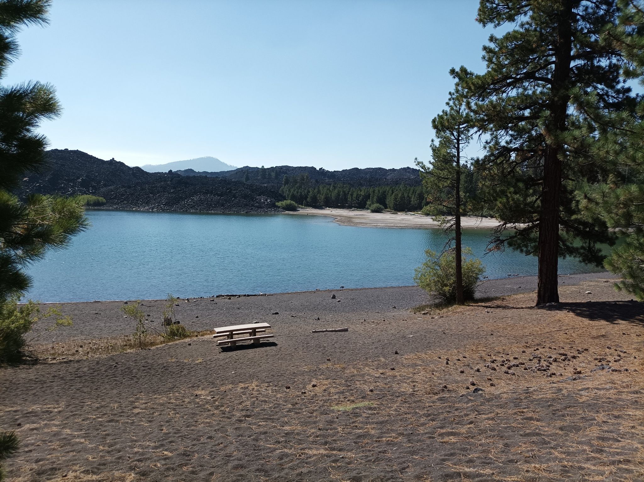A picnic table on a sandy shore next to a calm lake, surrounded by trees. - Butte Lake loop from Butte Lake Parking