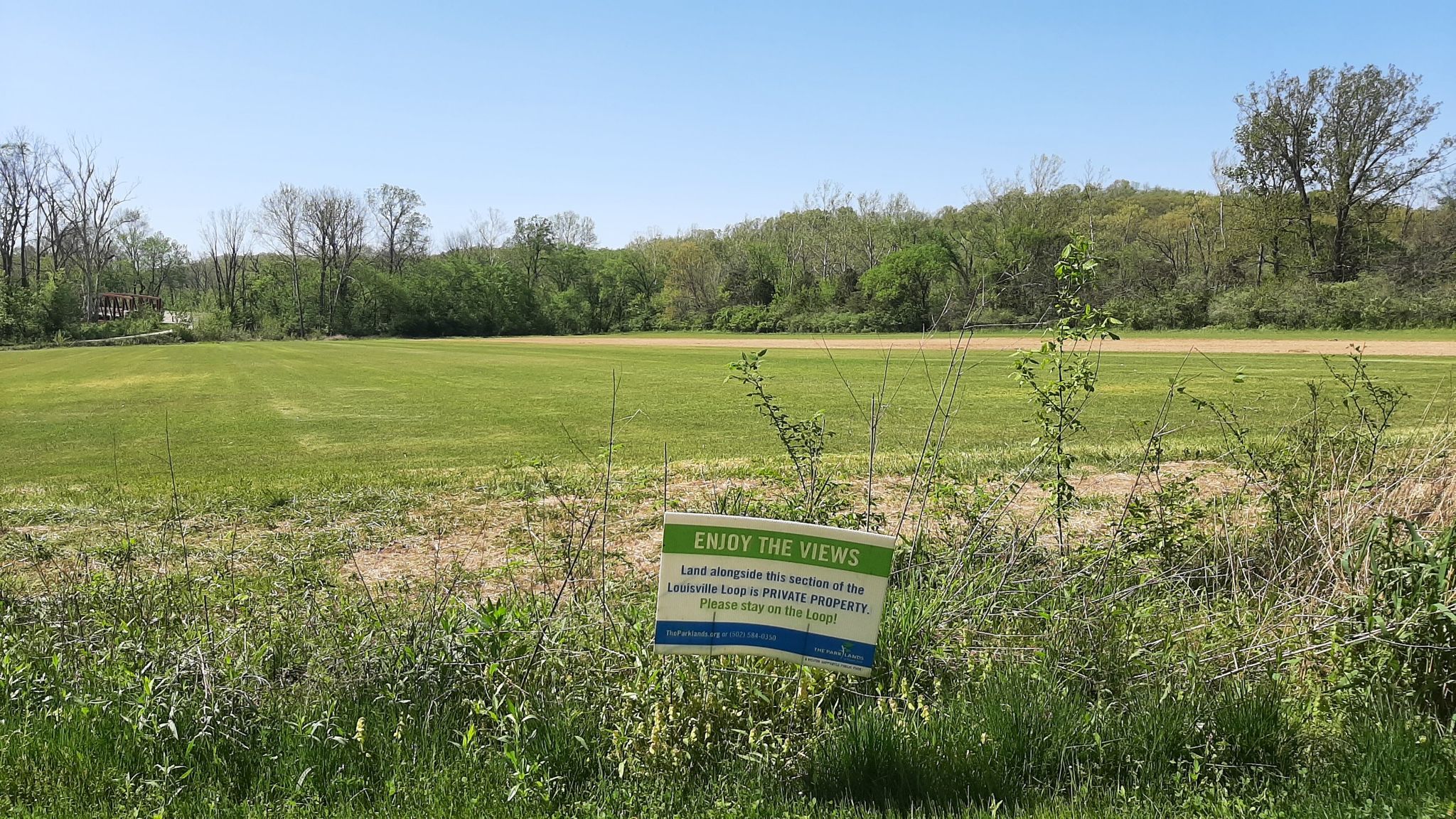 A sign in a sunny, expansive green field with distant trees. - Louisville Loop Trail