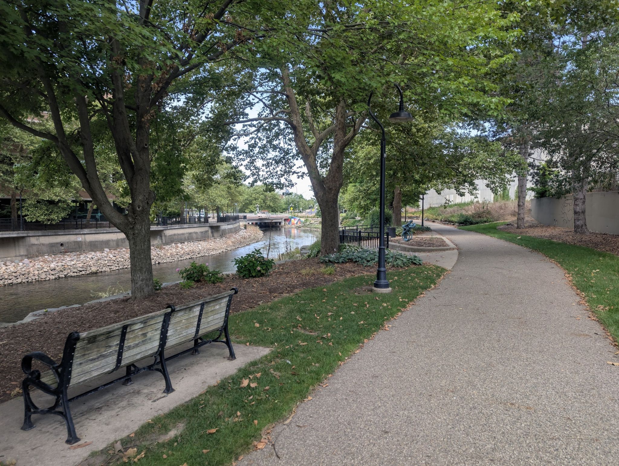 Paved path next to a rocky stream, with benches and trees. - Kalamazoo River loop from Kellogg Community College