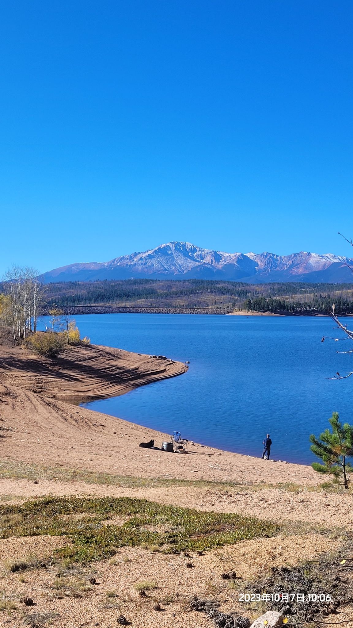 Blue lake with sandy shore, distant snow-capped mountains, and a person standing. - Rampart Reservoir loop from Woodland Park