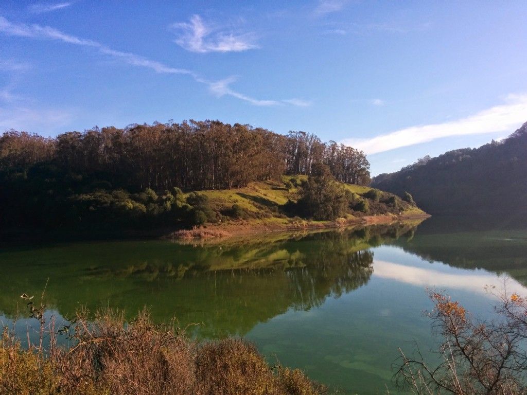 Greenish lake reflecting trees and sky, surrounded by hills on a sunny day. - Chabot Dam and Spillway – Great view of Lake Chabot loop from Lake Chabot Regional Park