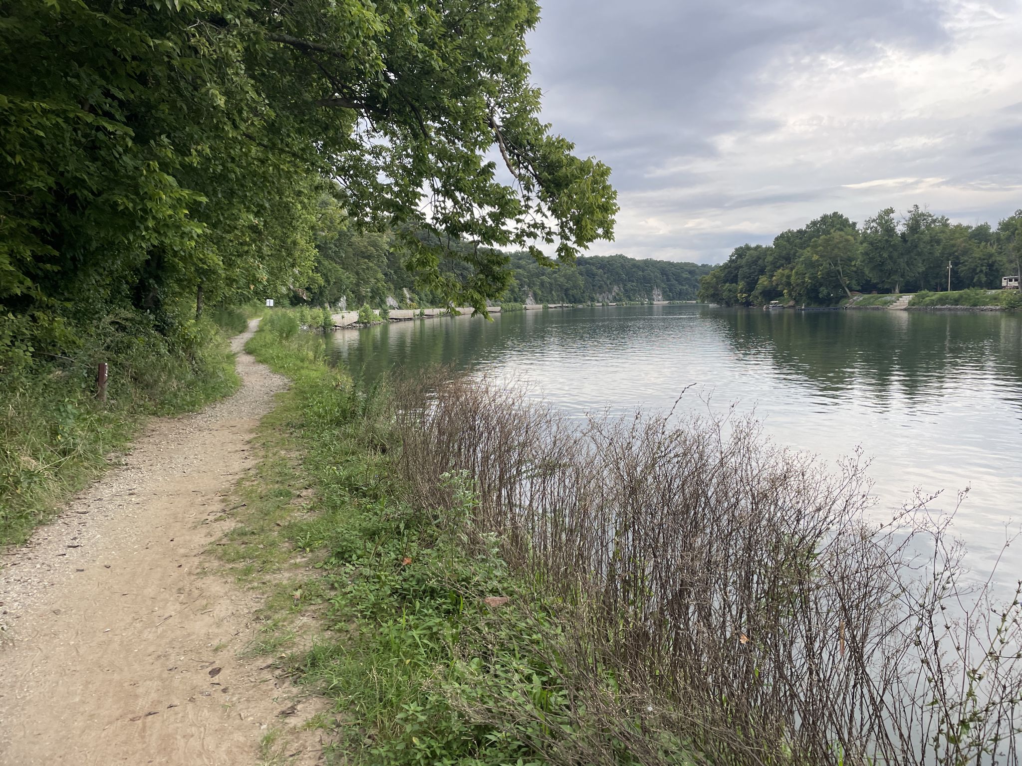 Dirt path alongside a calm river with lush green trees under a cloudy sky. - Taylors Landing to McMahon's Mill via C and O Canal Trail