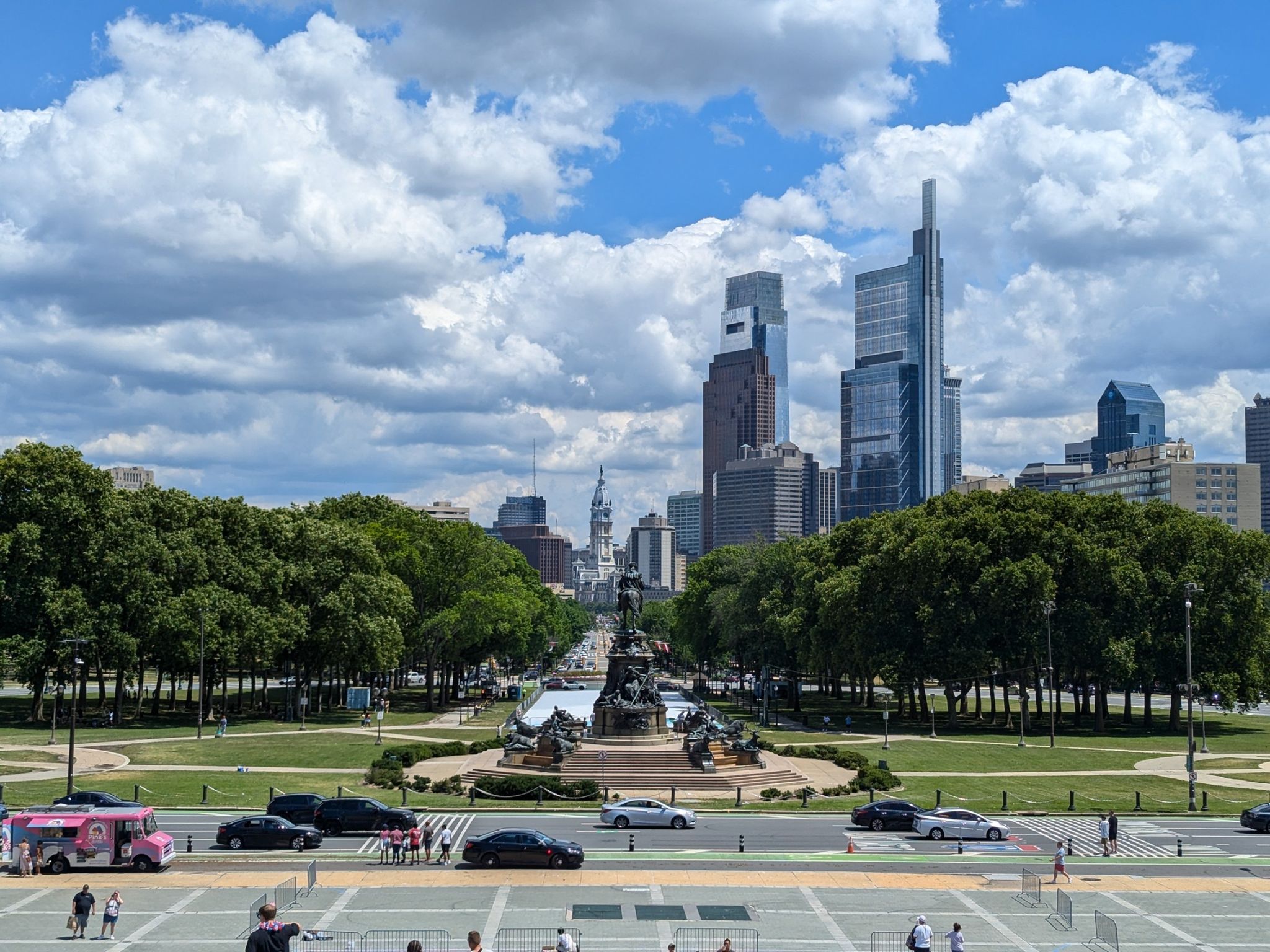 Distant city skyline beyond a park, monument, and busy street with people. - Shakespeare Memorial – Rocky Statue loop from Philadelphia