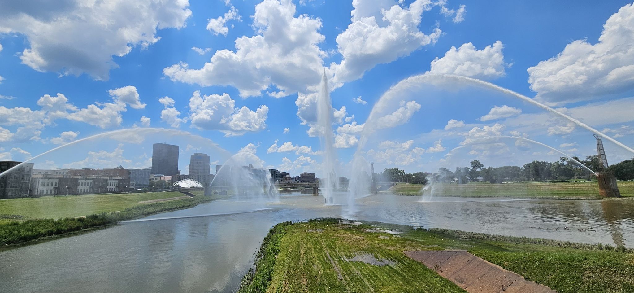 Multiple large fountains spraying water high from a river on a sunny day. - Mad River Recreation Trail