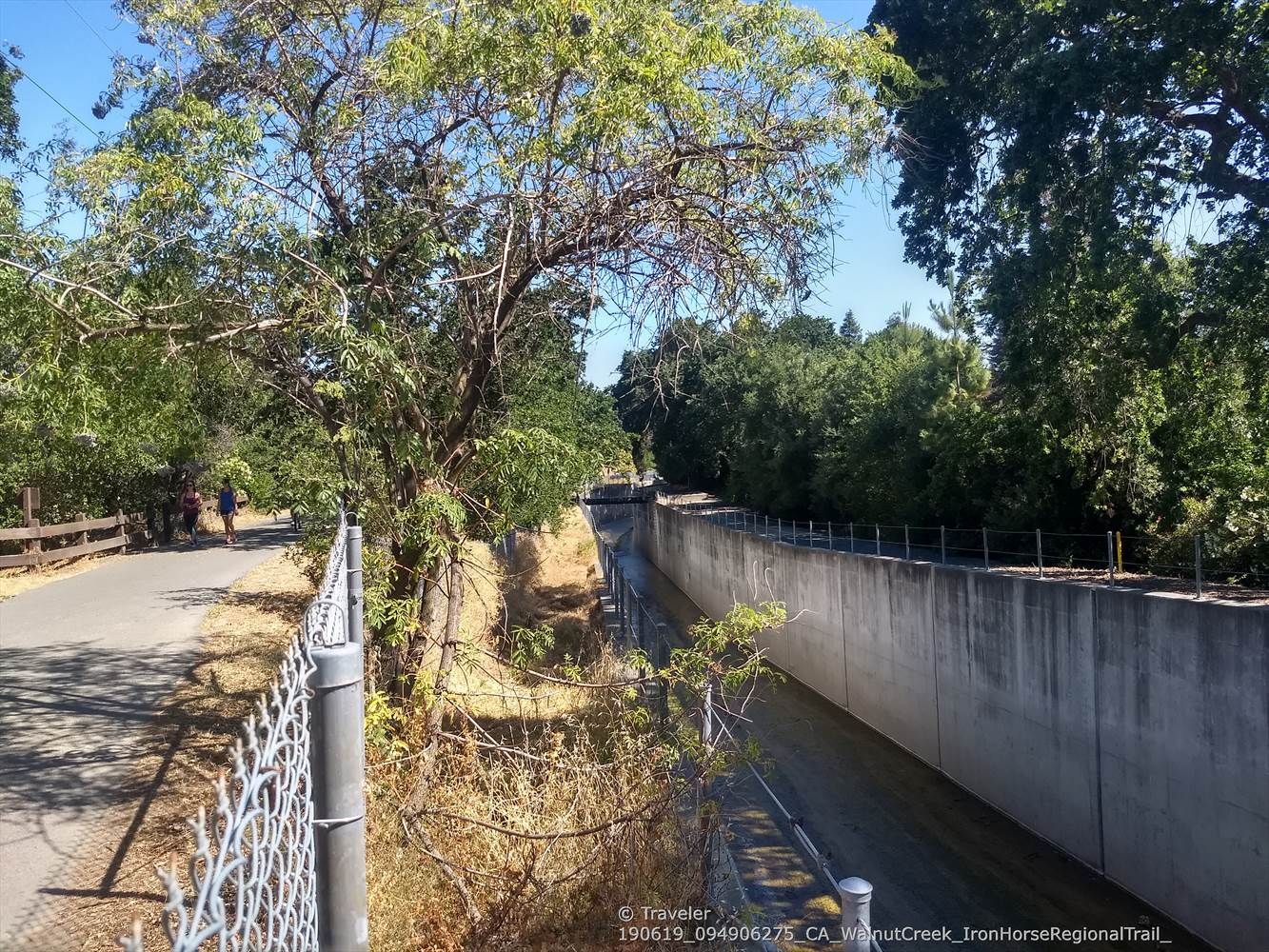 Paved path alongside a concrete channel with dense green trees. People walking. - Iron Horse Regional Trail