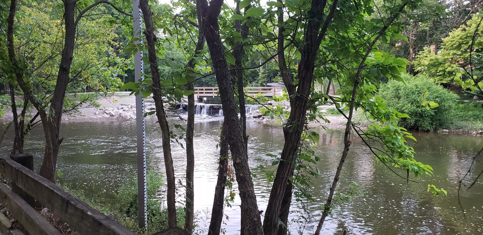 A small waterfall and wooden bridge seen through green trees by a river. - Waterfall at Creekside Park – Fountain at Creekside Plaza loop from Techcenter Dr & AEP