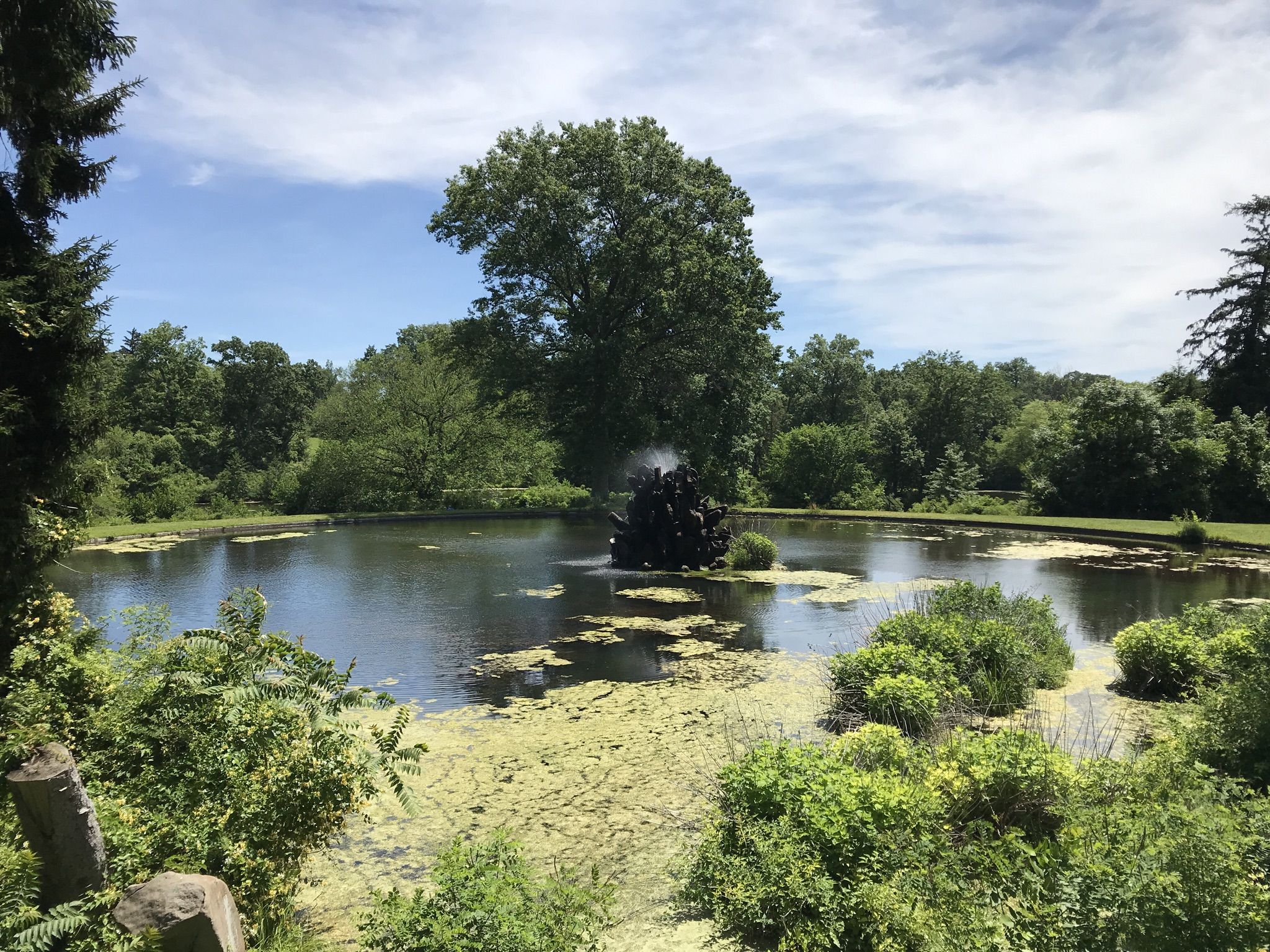 A pond with a dark fountain, surrounded by lush green trees on a sunny day. - Great Falls loop from Manville