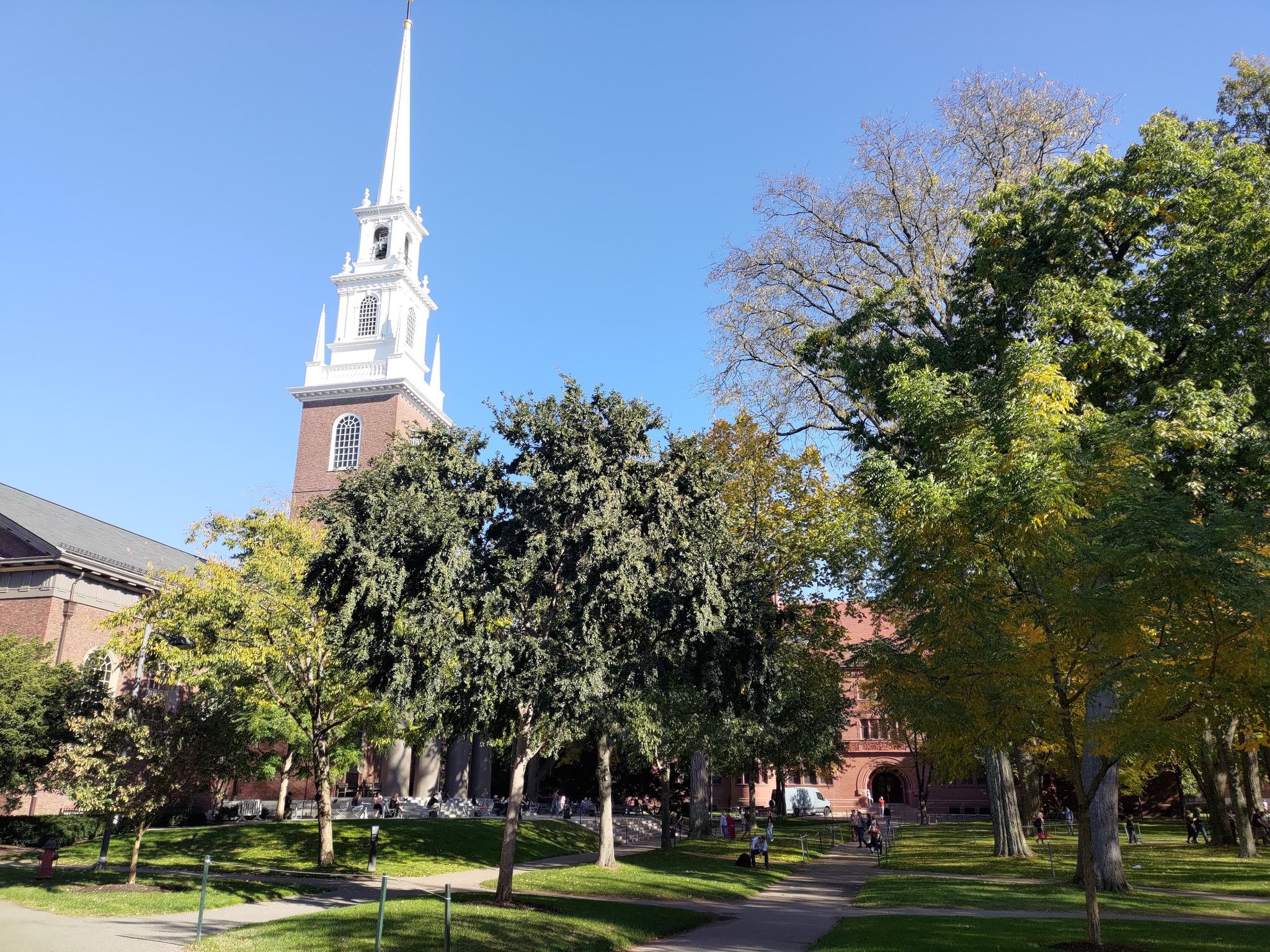 A tall white steeple rises above green trees and a brick building. - Harvard Yard Tour
