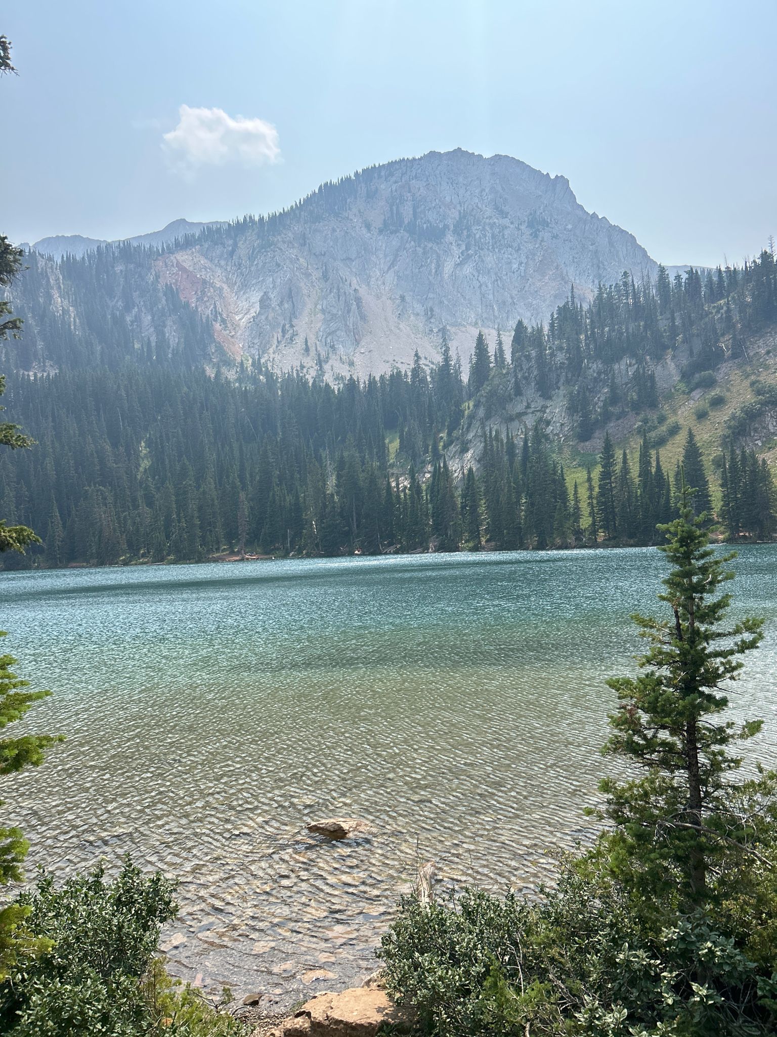 Clear mountain lake with pine trees and a large rocky peak under hazy sky. - Fairy Lake loop