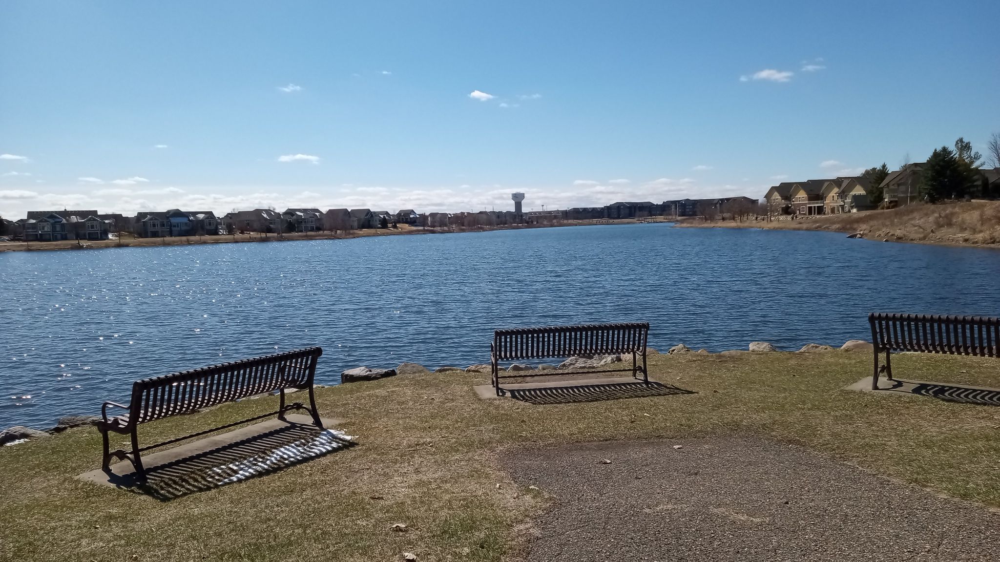 Three empty benches overlooking a wide lake with distant houses under blue sky. - Cobblestone Lake loop from Claret Springs East