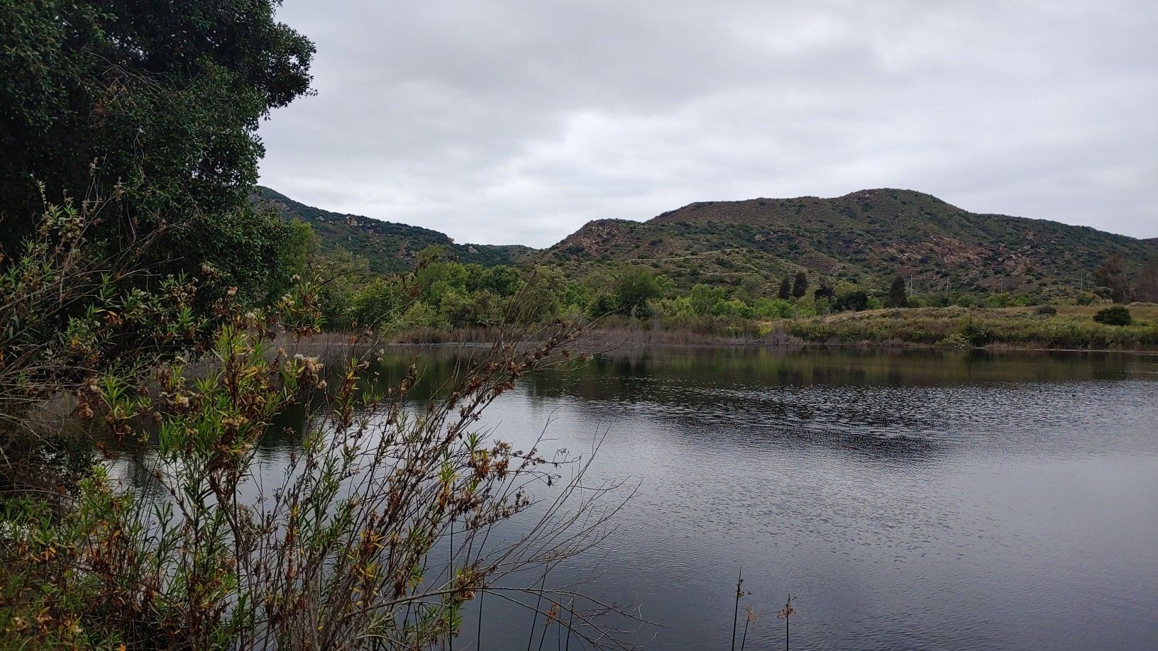 A calm lake surrounded by green trees and hills under a cloudy sky. - Barbara's Lake Loop Trail