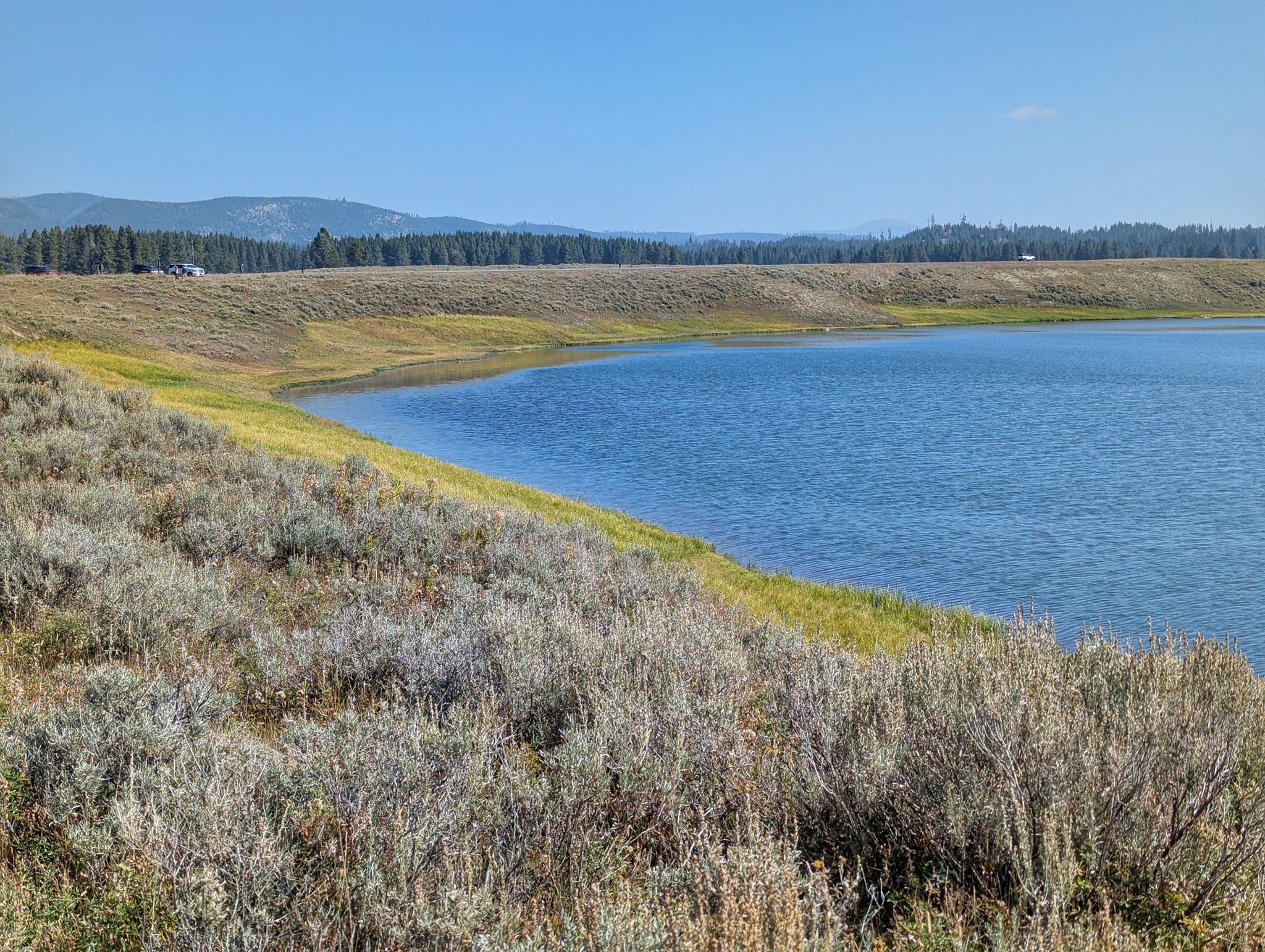 Blue lake with grassy shores, distant hills, and clear sky. - Storm Point Trail