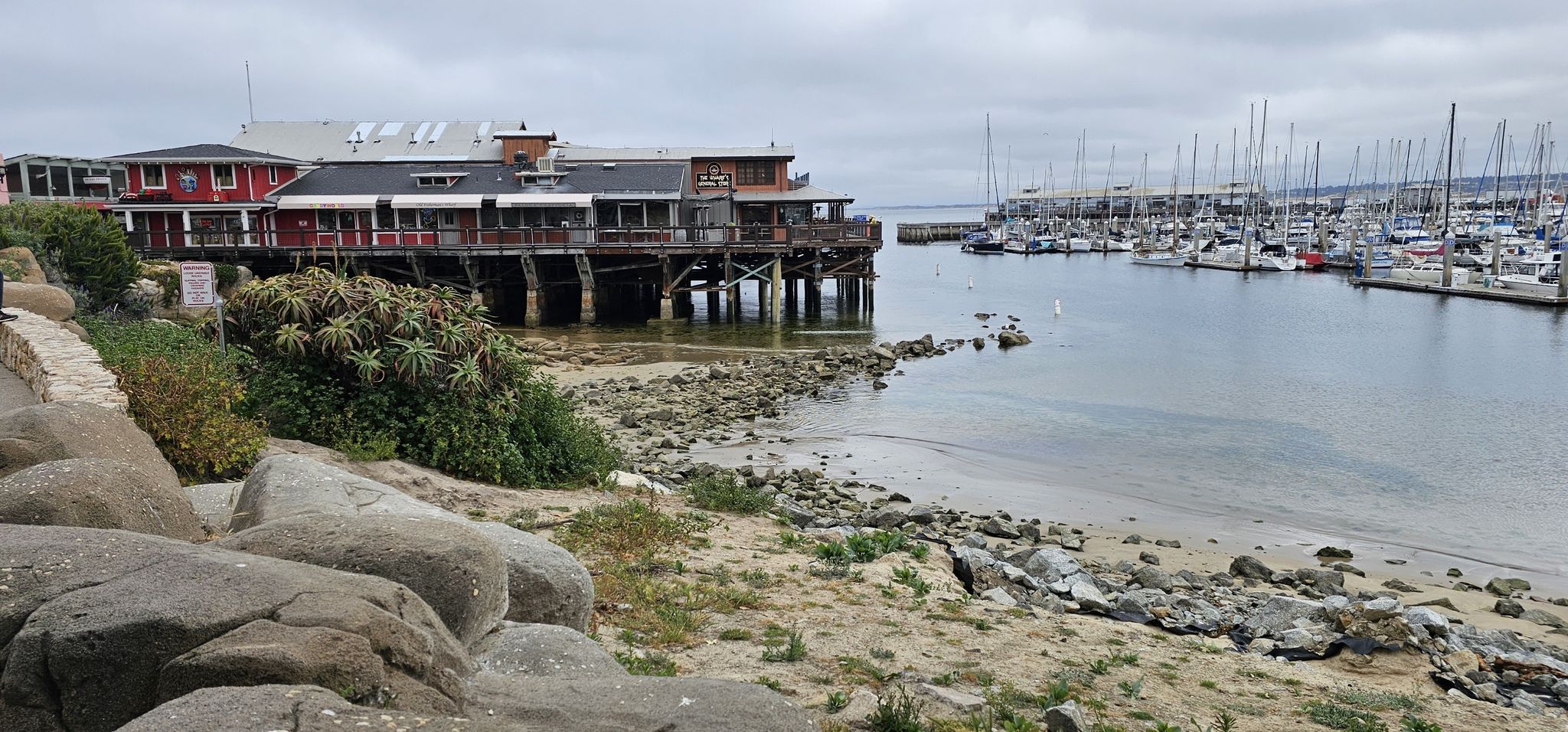 Pier buildings over water, boats in a harbor, and a rocky shore on a cloudy day. - Fishermans Wharf – Monterey State Beach loop from Monterey State Beach
