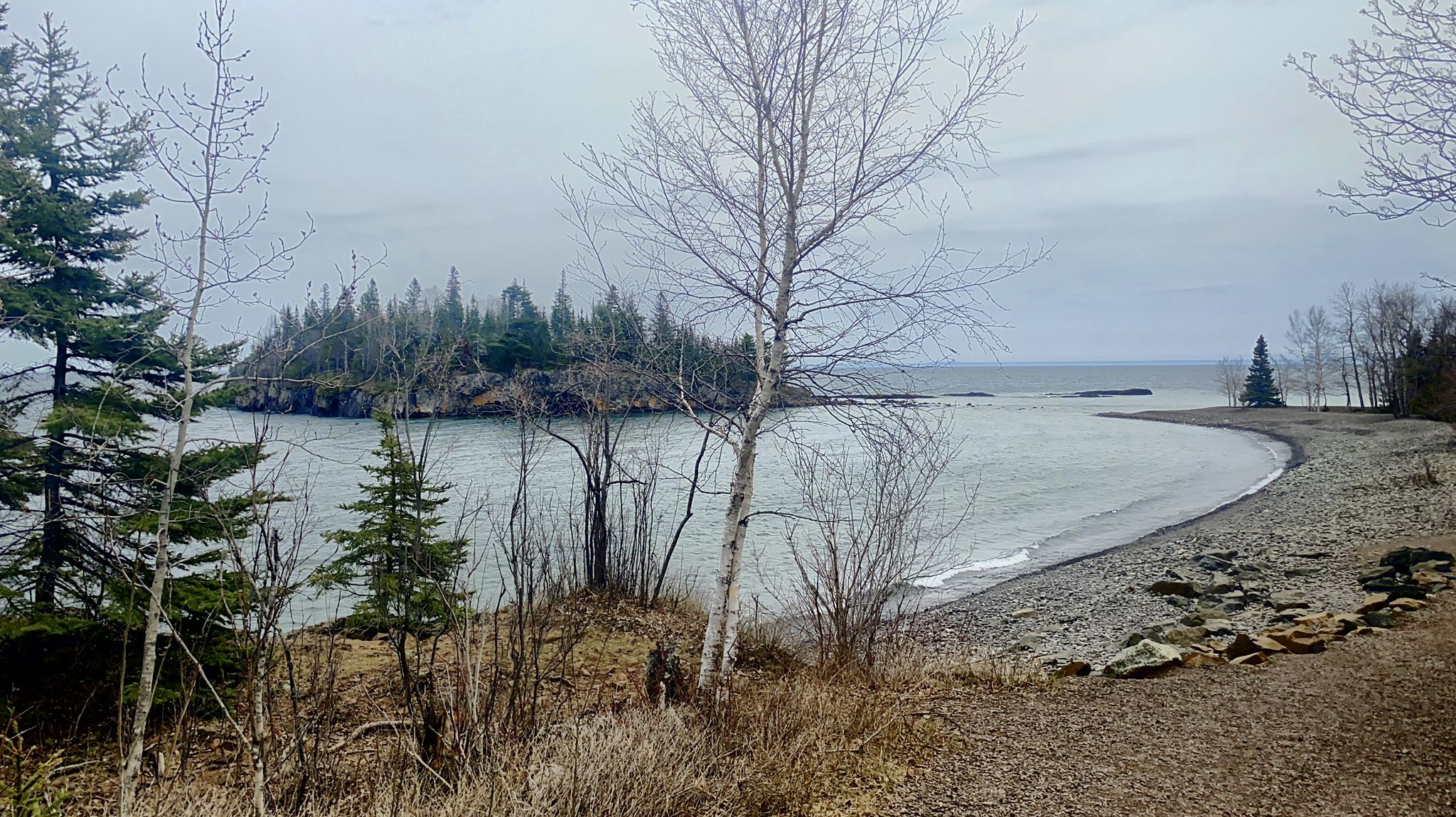 Overcast view of a rocky beach, calm water, and a forested island. - Split Rock Lighthouse – View of Split Rock Lighthouse loop from Split Rock Lighthouse