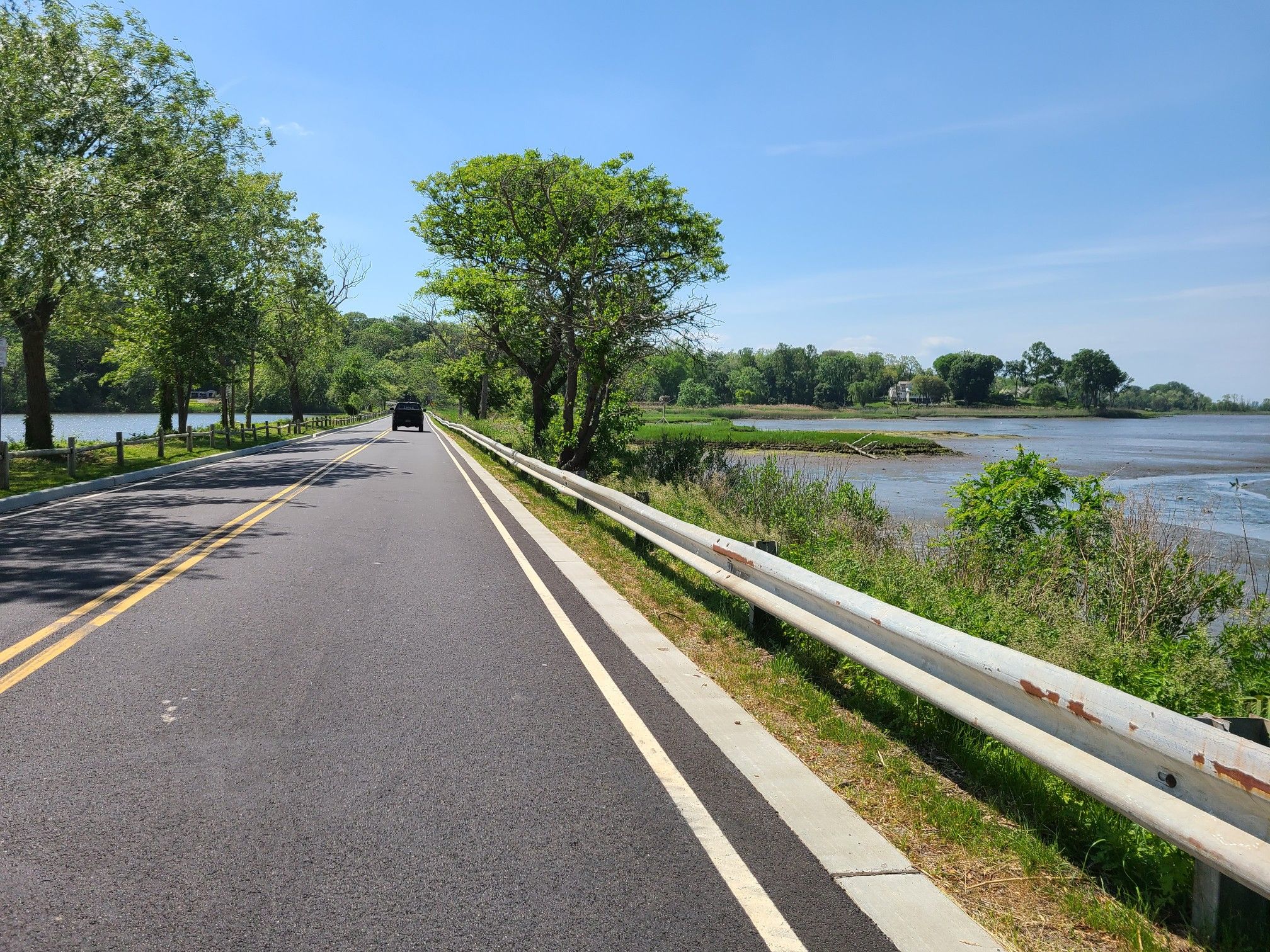 Sunny road alongside a calm body of water with a distant car. - Muttontown Road – Welwyn Preserve County Park loop from Hicksville