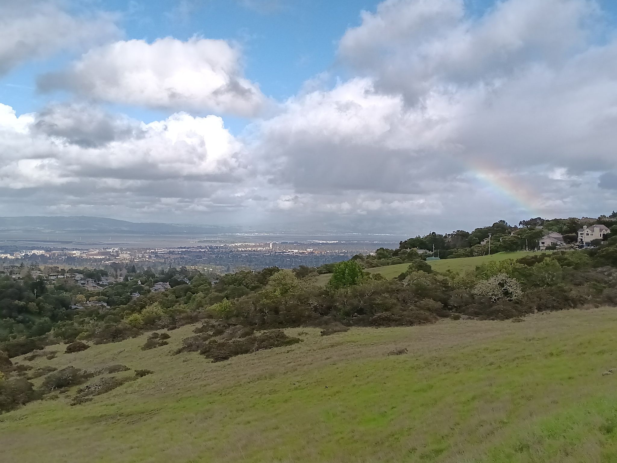 A wide landscape view of a city below hills, with a rainbow. - Edgewood Trail loop from Emerald Lake Hills