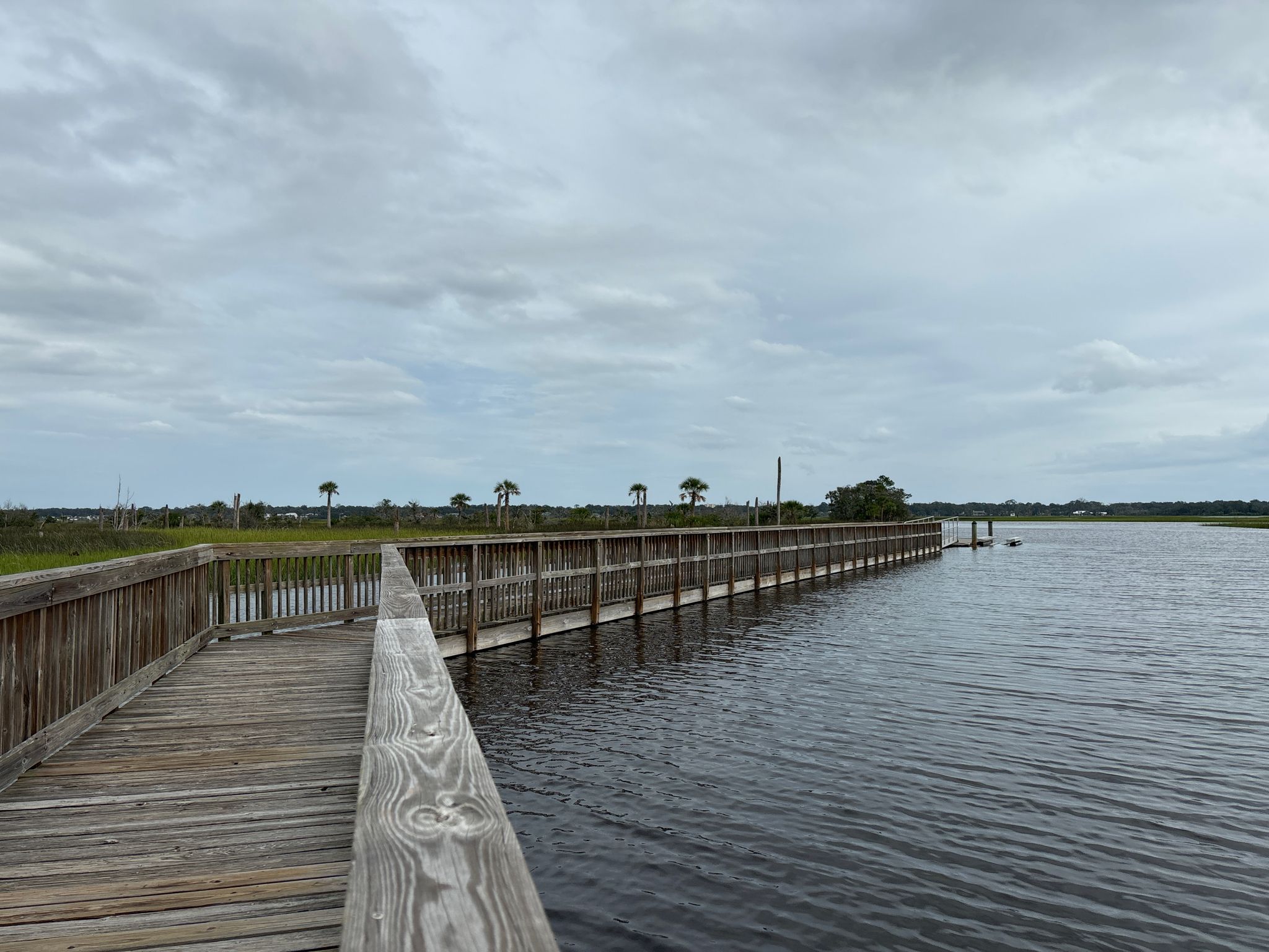A long wooden boardwalk extends over dark water, with marshland in the distance under a cloudy sky. - Island Trail