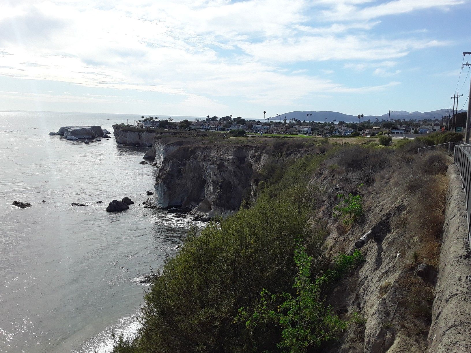 Rocky ocean cliffs and a distant town under a partly cloudy sky. - Pismo Beach – Avila Beach boardwalk ⛱️ loop from San Luis Obispo