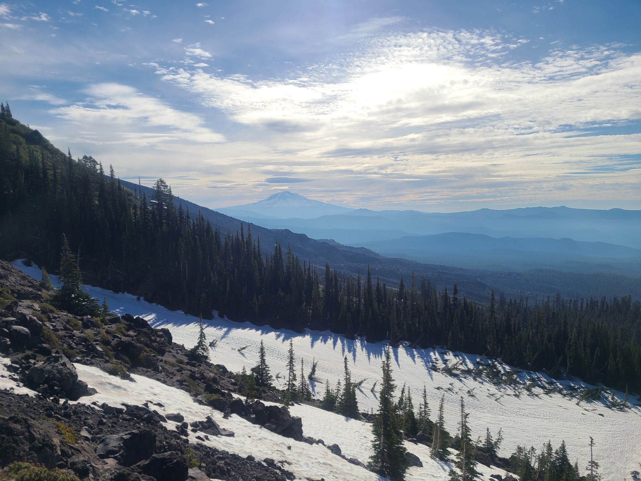 Snow patches on a forested mountain slope with a distant peak under cloudy sky. - Mount Saint Helens Summit via Ptarmigan Trail
