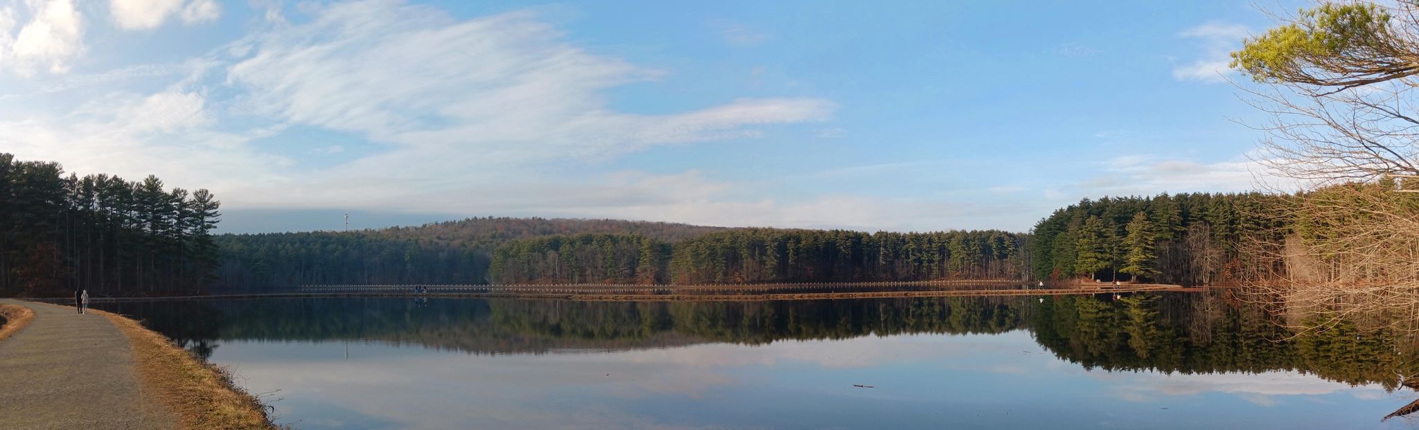 Panoramic view of a lake reflecting forested hills, with a person walking. - Ashley Reservoir Trail