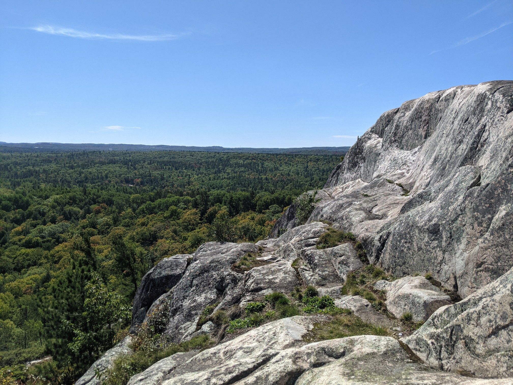 Steep gray rock formations overlook a vast green forest under a clear sky. - Hogback Mountain