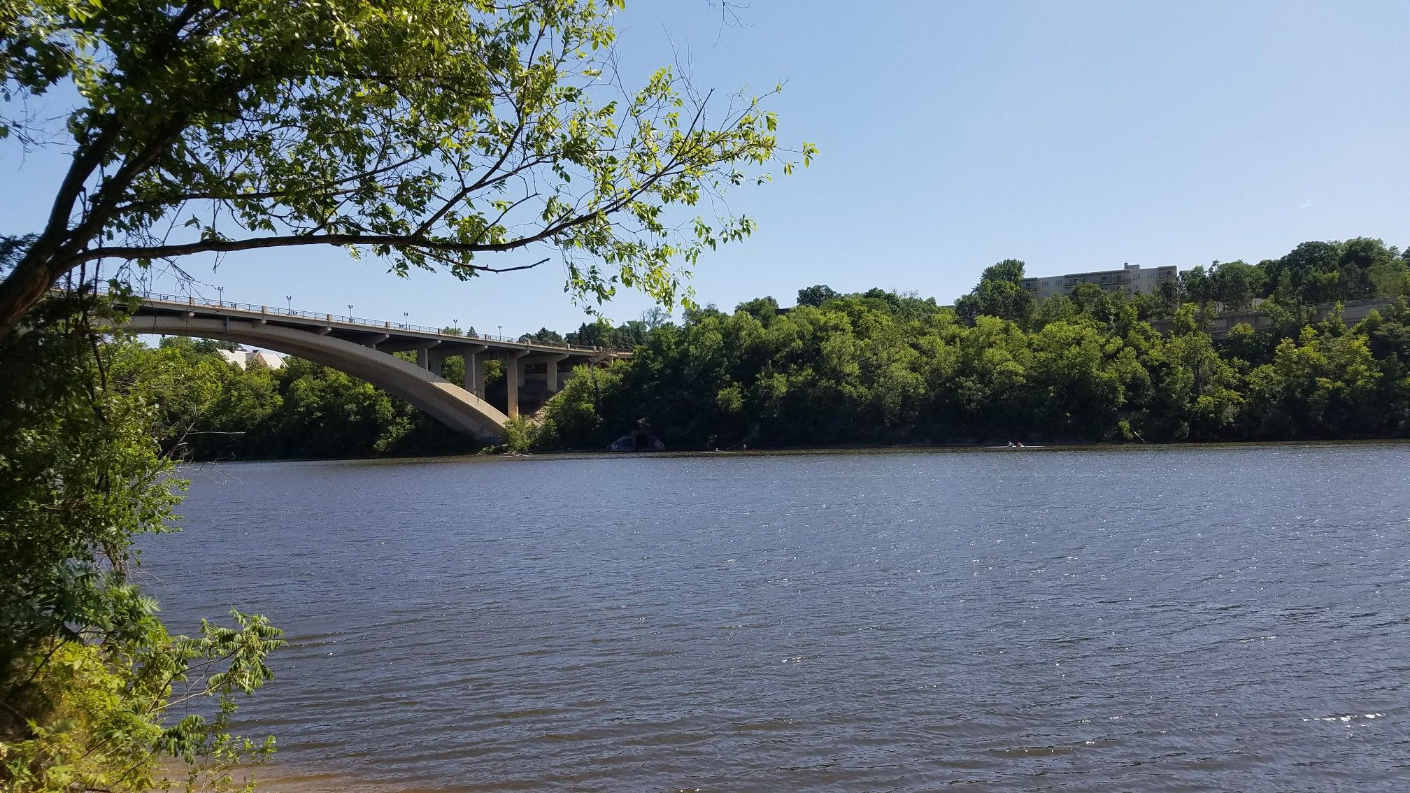 Wide river with an arched bridge and green trees under a bright sky. - Winchell Trail