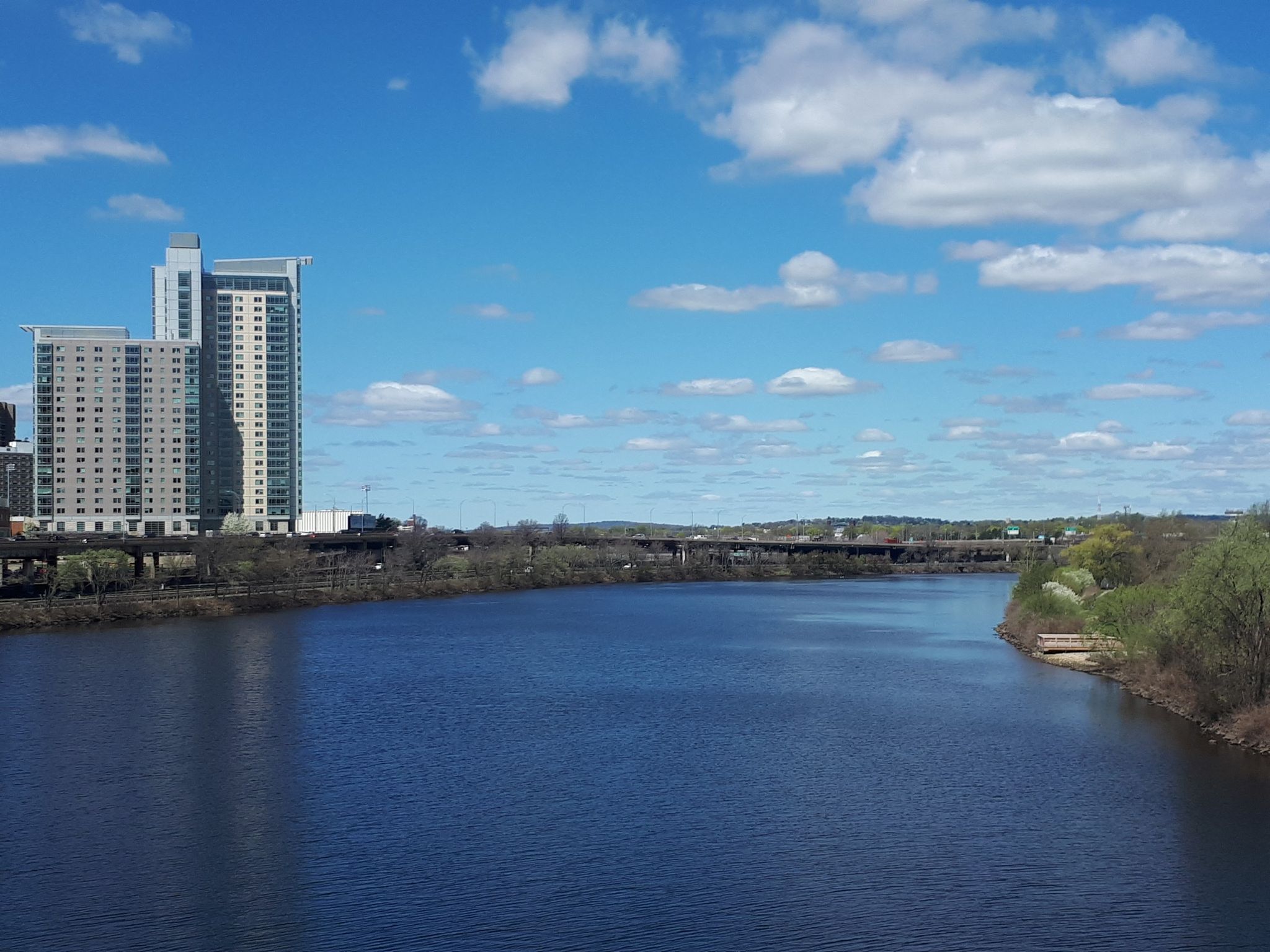 Wide river, tall buildings, and distant bridge under a bright blue sky. - Charles River Southwest Corridor Trail