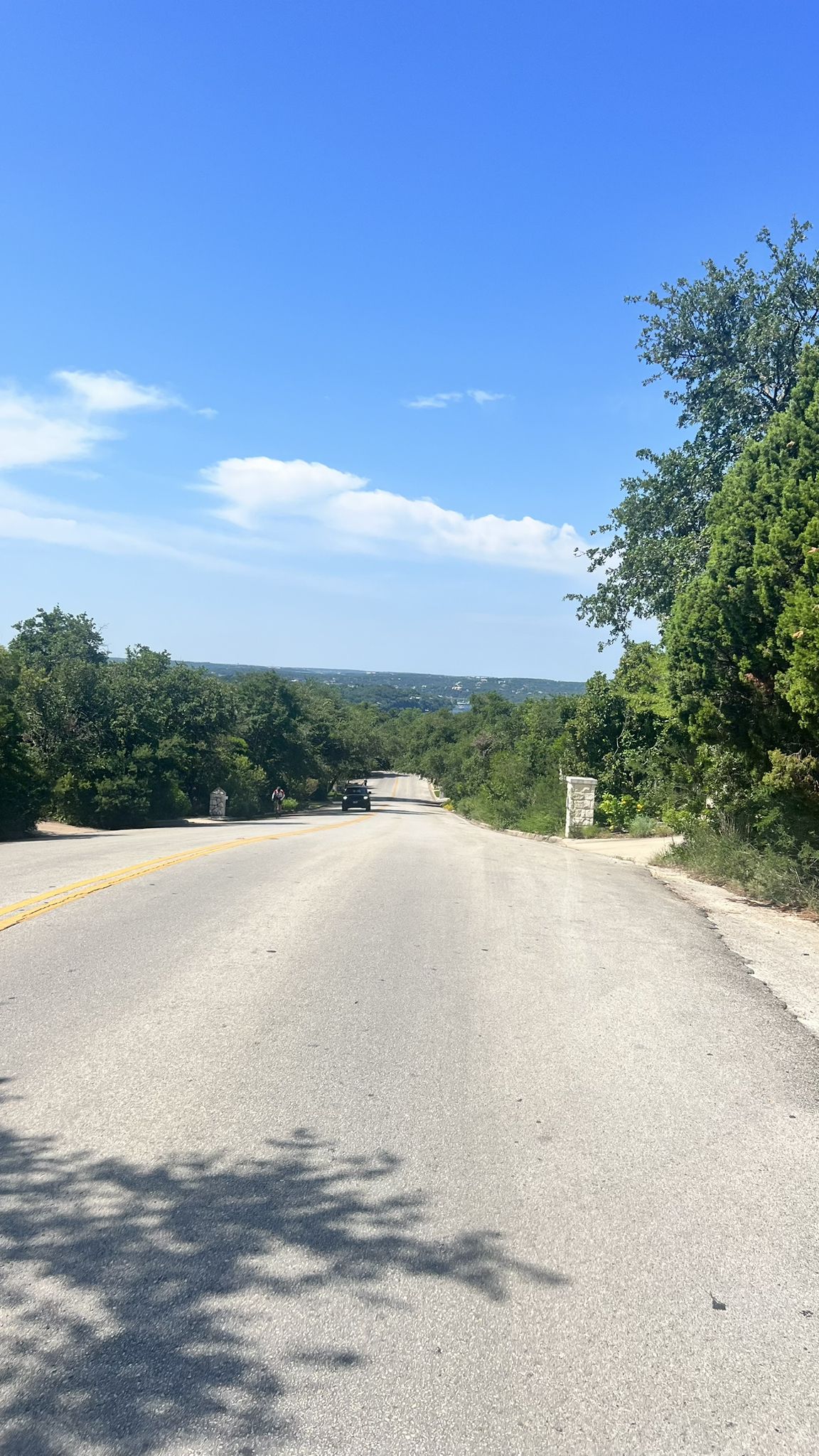 Empty road descends through lush green trees under a bright blue sky. - River Place Nature Reserve & Lake Austin loop