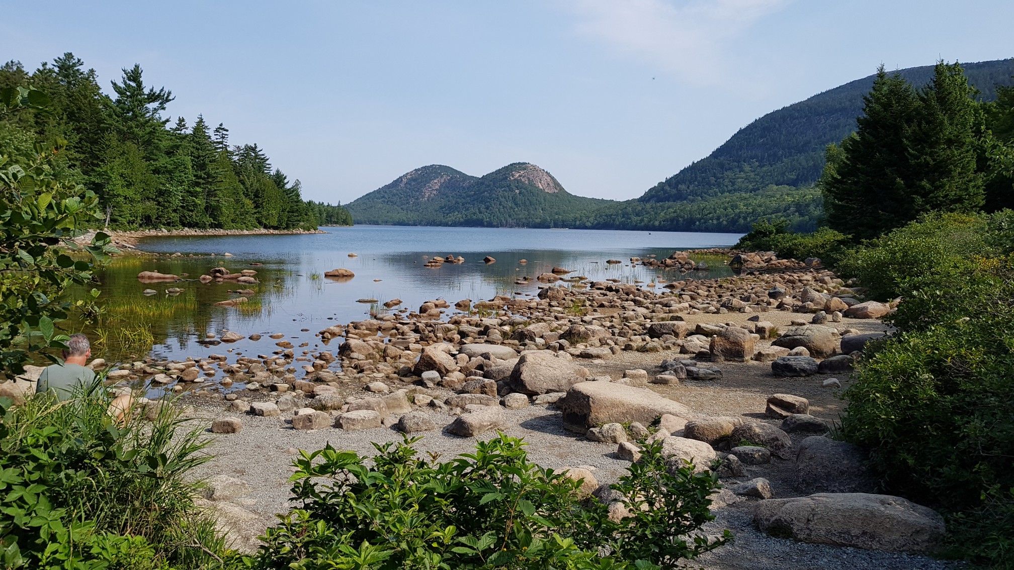 Rocky lake shore with distant mountains, a person on the left, sunny. - Pemetic Mountain Loop