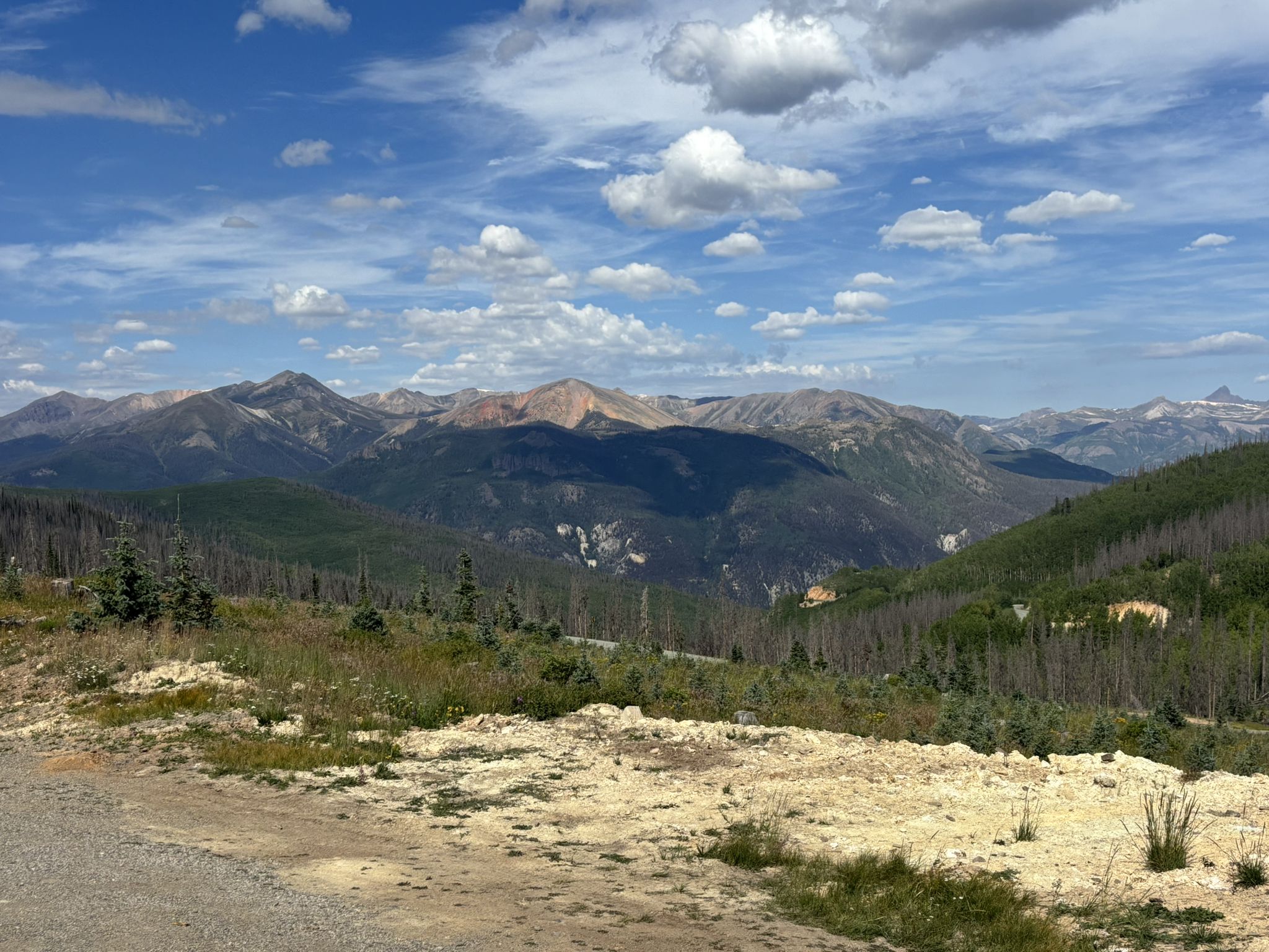 Vast mountain range under a partly cloudy sky, with forested slopes and a dirt foreground. - Slumgullion Pass — Lake City