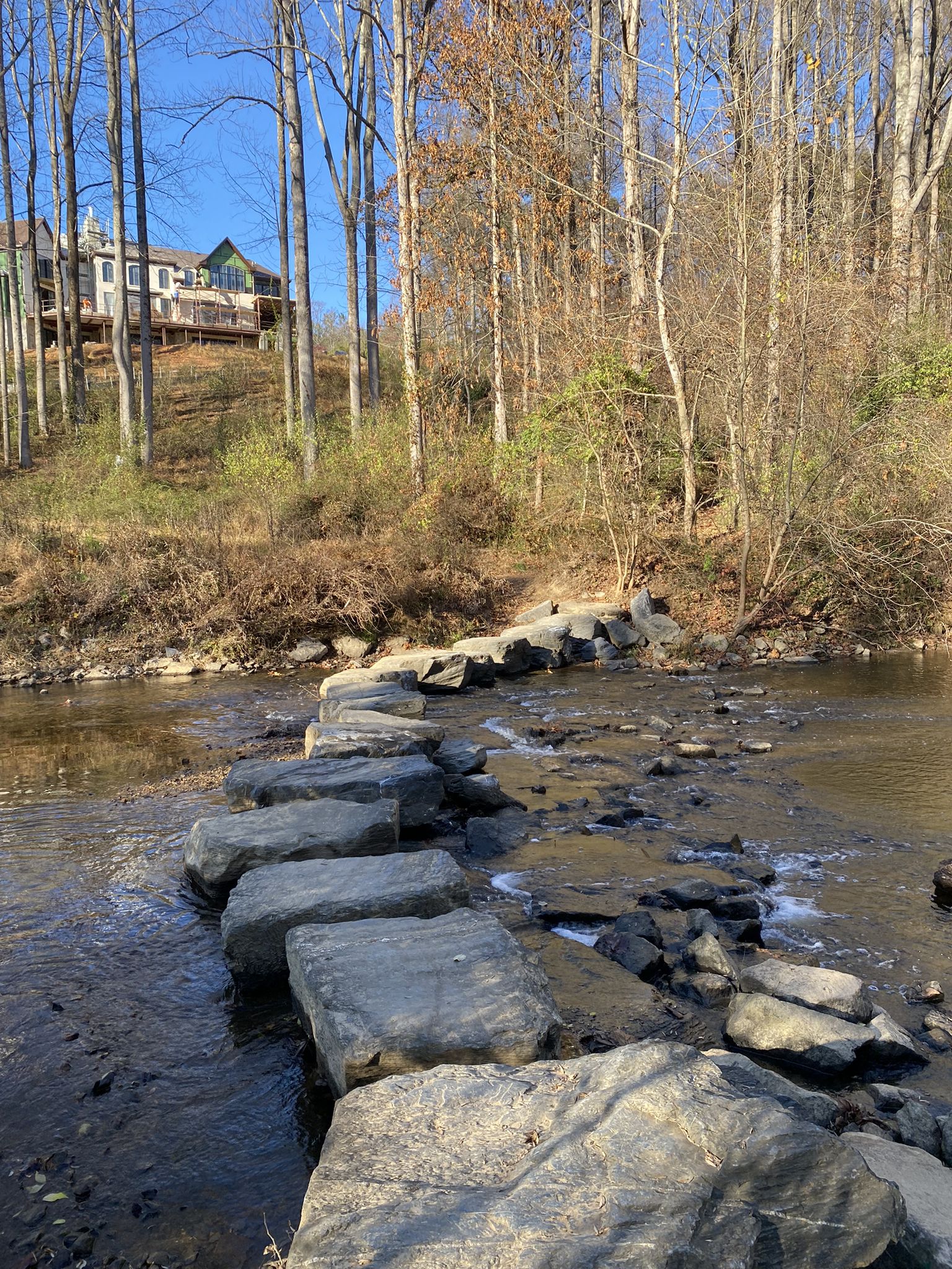 Large stepping stones cross a shallow river, bordered by bare trees and a house. - Cross Country Trail loop from Chesapeake and Ohio Canal National Historical Park
